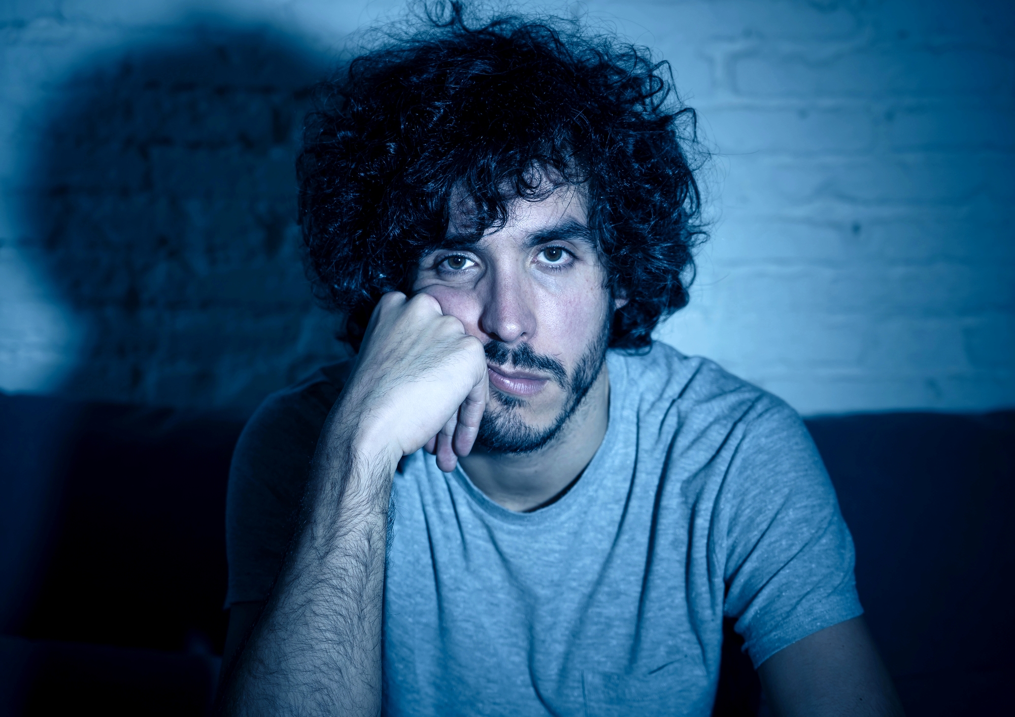 A man with curly hair and a beard leans his head on his hand while sitting against a light-colored brick wall. He is wearing a gray t-shirt and appears to be in a dimly lit setting, highlighted by a blue-toned light casting a shadow behind him.