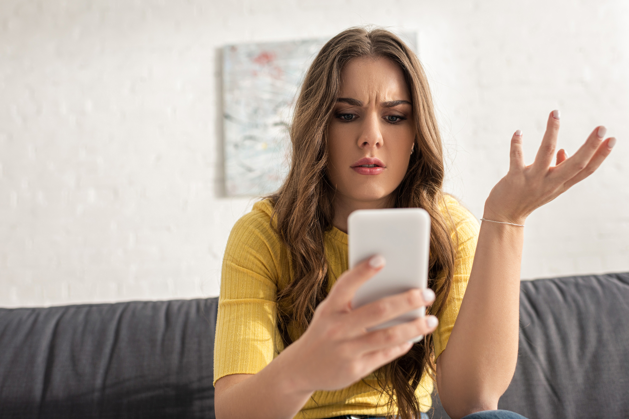 A young woman with long brown hair, wearing a yellow shirt, sits on a grey couch. She holds a smartphone in one hand and looks at the screen with a confused and frustrated expression, her other hand raised in a questioning gesture. A painting is blurred in the background.