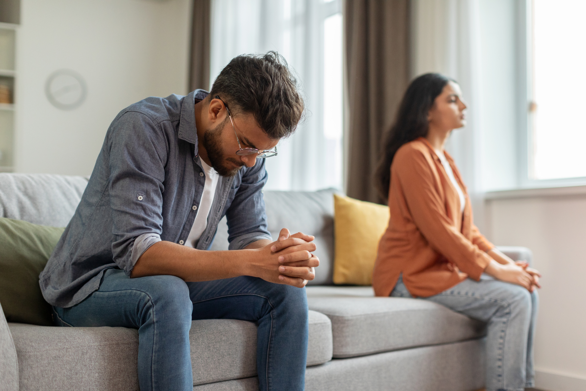 A man and woman sit on a couch, facing away from each other in a tense atmosphere. The man, wearing glasses and a denim shirt, sits with his head down and hands clasped in front of him. The woman, in an orange shirt, sits upright with her back turned to the man.