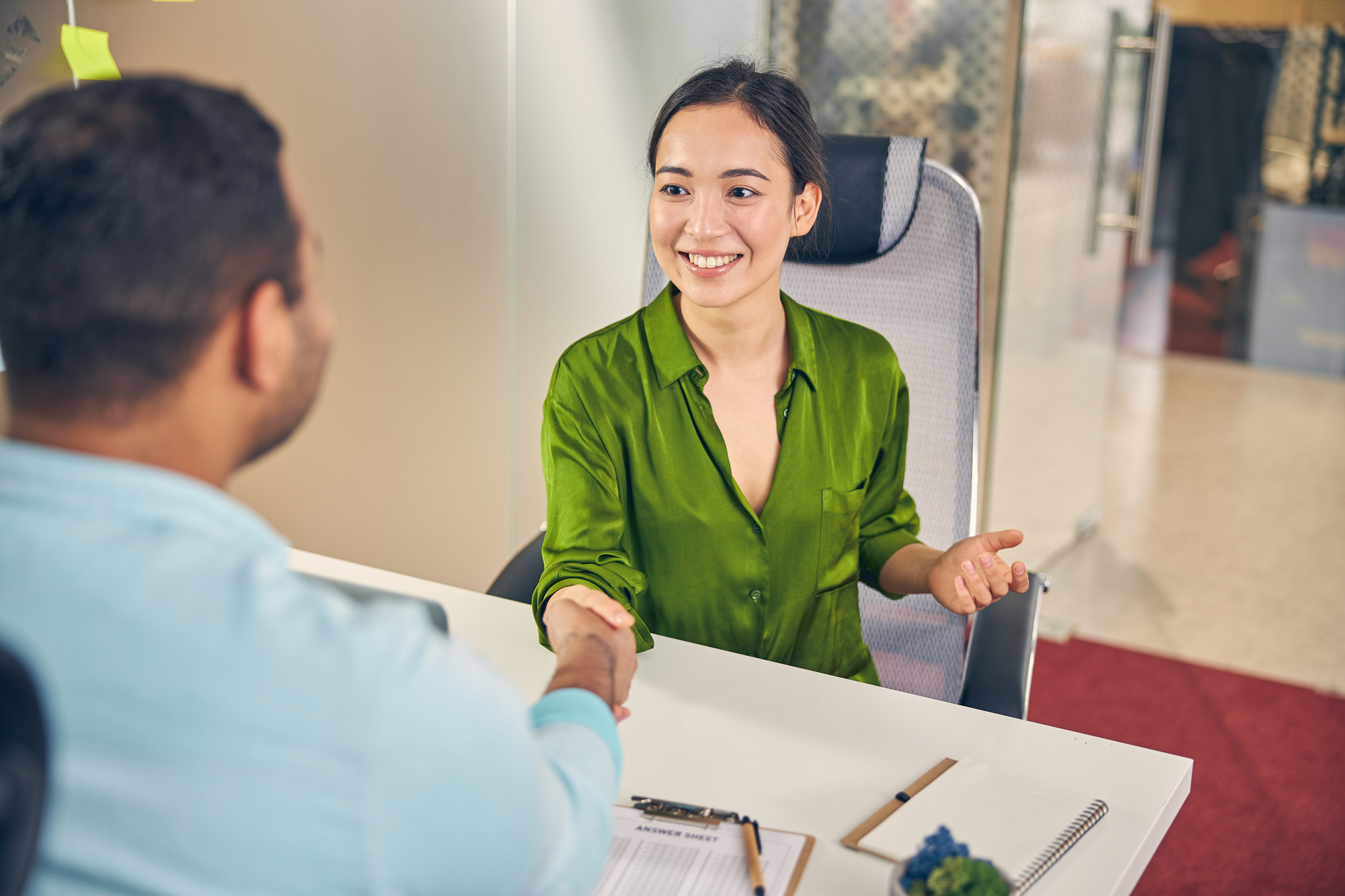 A woman in a green shirt sits at a table, smiling, and shakes hands with a man whose back is to the camera. A clipboard, notepad, and pen are on the white table in front of her. The background shows an office environment.