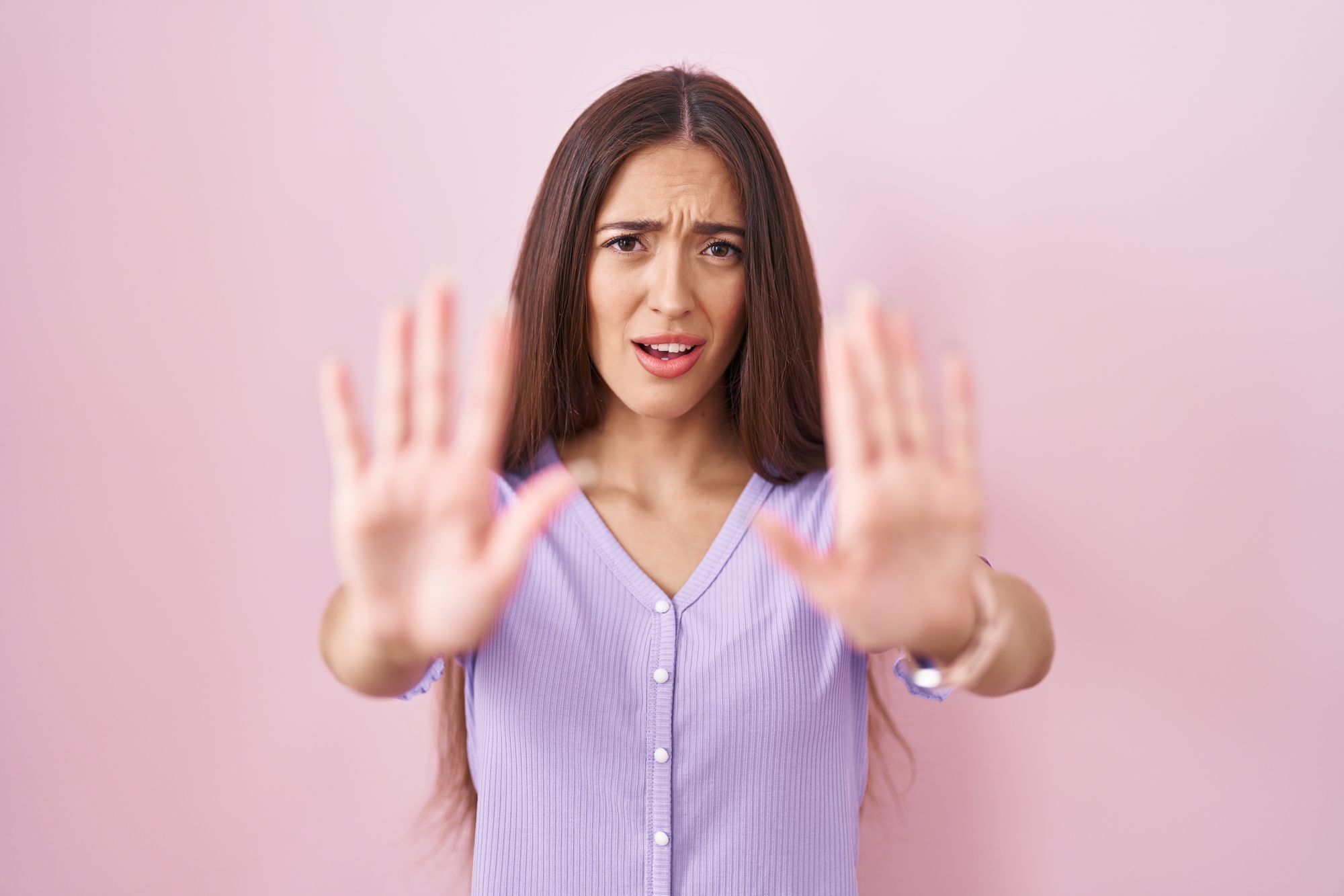 A woman with long brown hair is standing against a pink background, looking concerned. She is wearing a lavender top and extending both hands out in front of her as if to signal 'stop' or 'stay back.'.