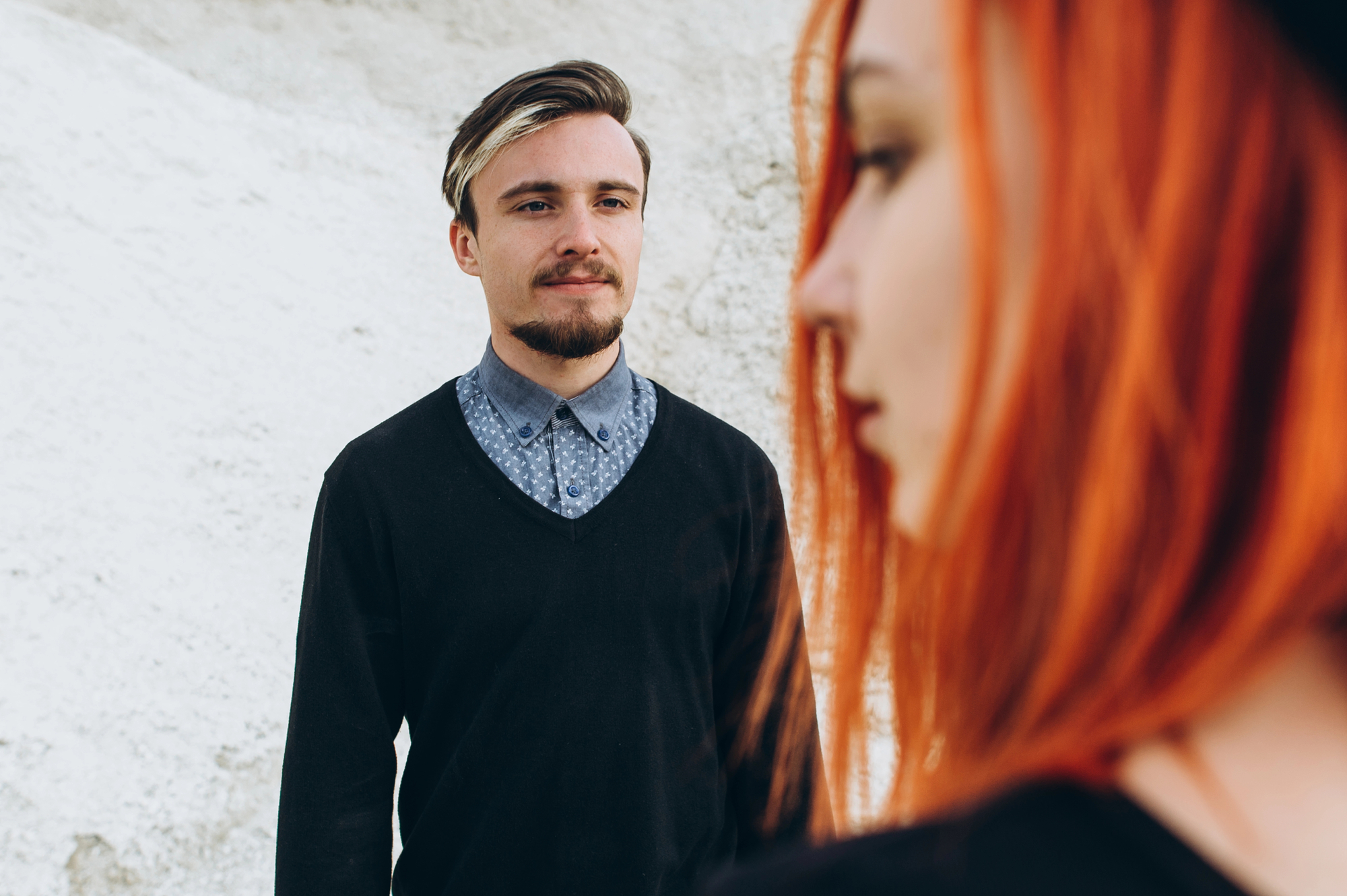 A man with short hair and a beard, wearing a black sweater over a blue patterned button-up shirt, is looking forward with a neutral expression. In the foreground, a woman with vibrant red hair, slightly out of focus, is partially visible in profile.