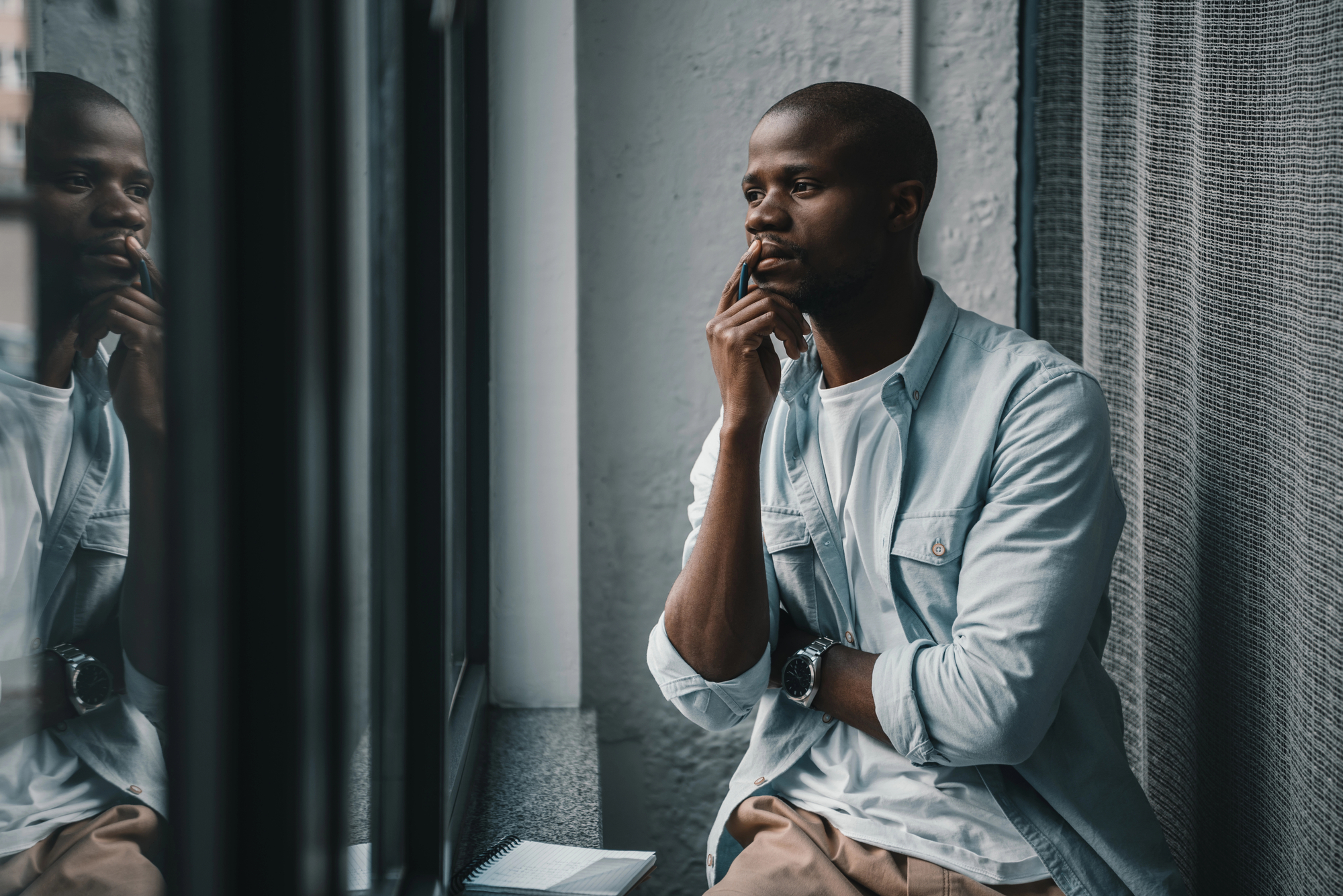 A man wearing a light denim shirt and a white t-shirt is sitting by a window, resting his chin on his hand, and looking thoughtfully outside. A notebook is on the window ledge next to him. The scene is calm and contemplative.