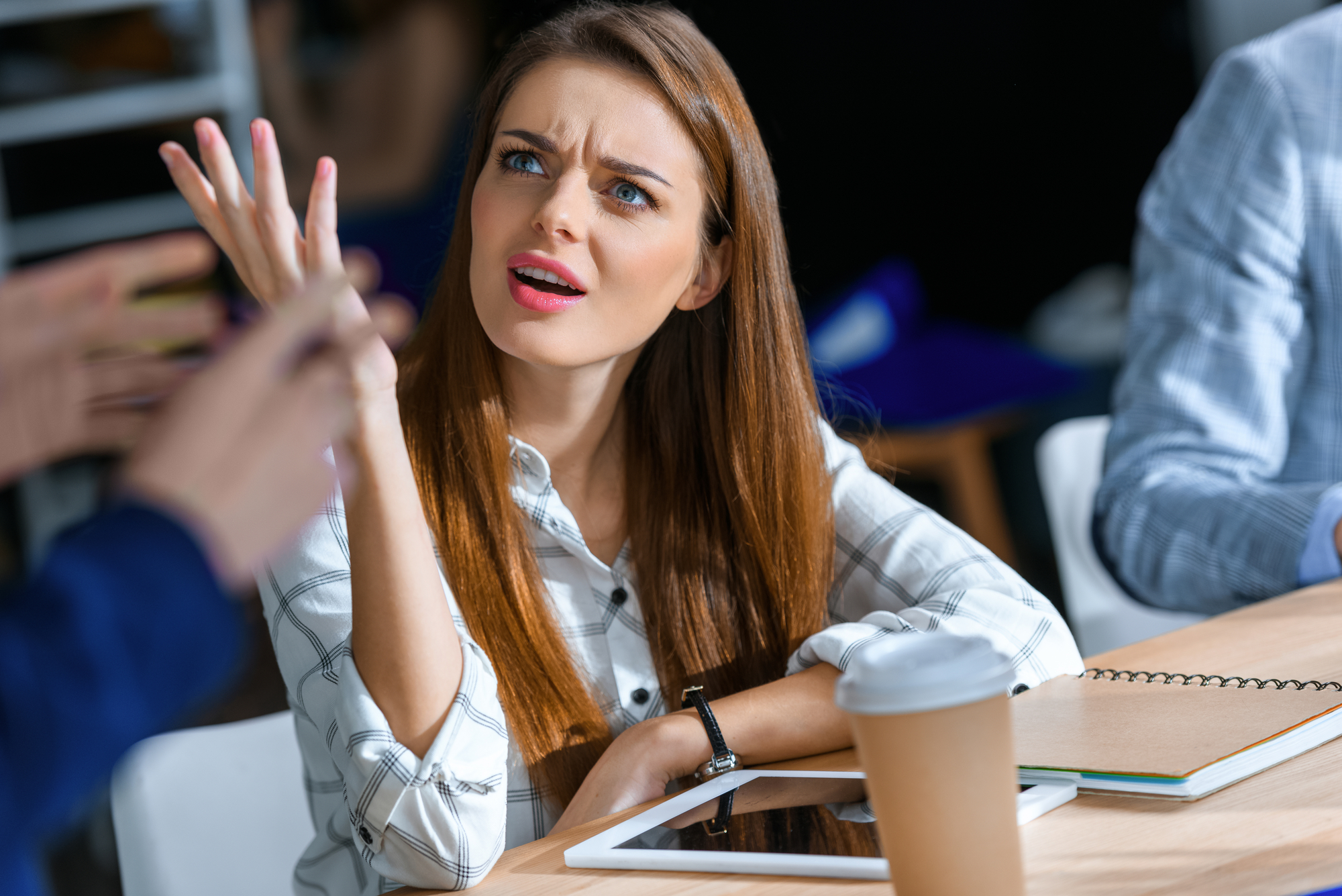 A woman with long brown hair and a white plaid shirt sits at a table, with her hand raised in a questioning gesture. An open notebook, a tablet, and a coffee cup are on the table in front of her. She appears to be engaged in a conversation with someone out of frame.