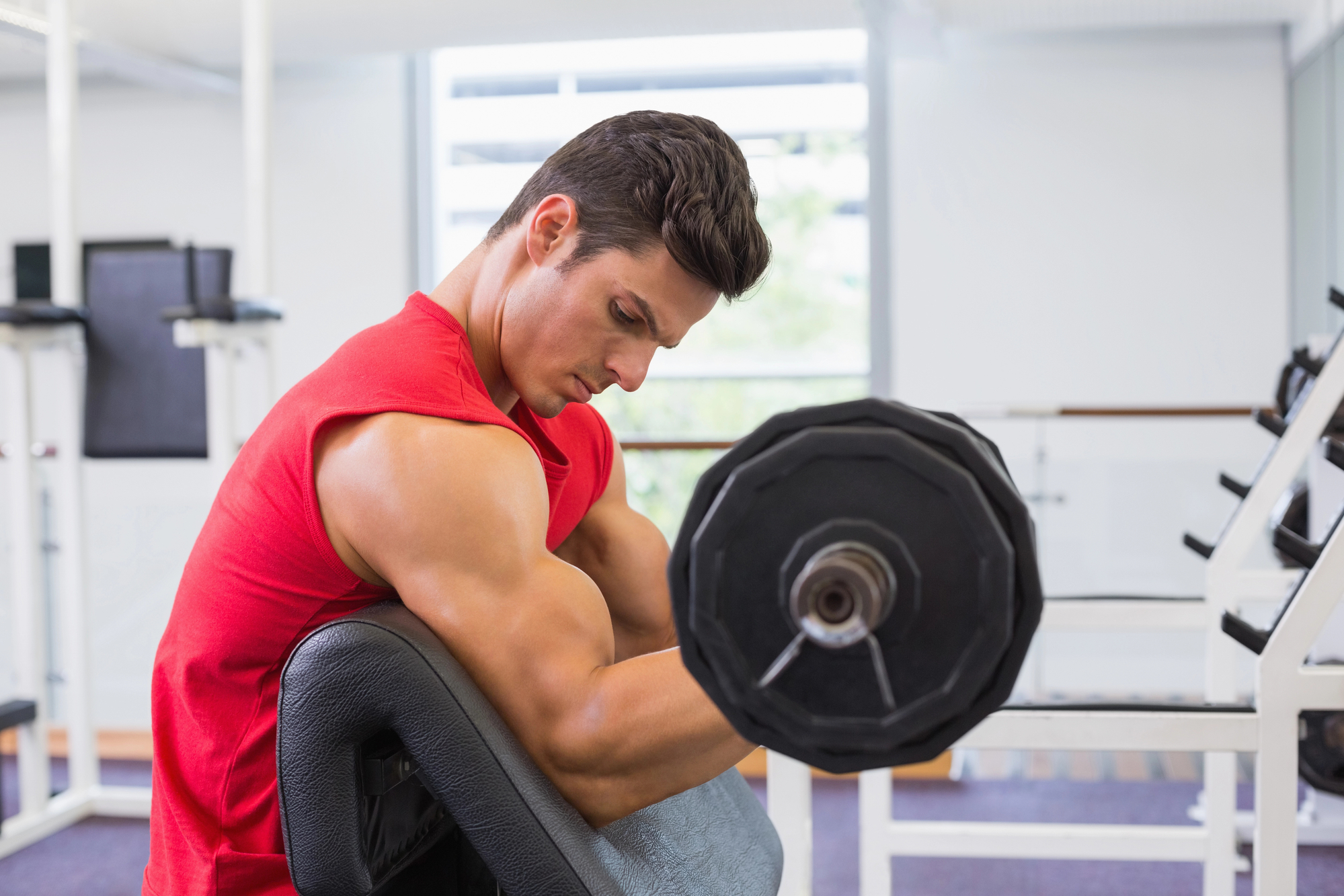 A man wearing a red sleeveless shirt is sitting on a preacher curl bench in a gym, lifting a barbell. His muscular arm is visible as he curls the weight, focused on his workout. The gym has various equipment and machines in the background.