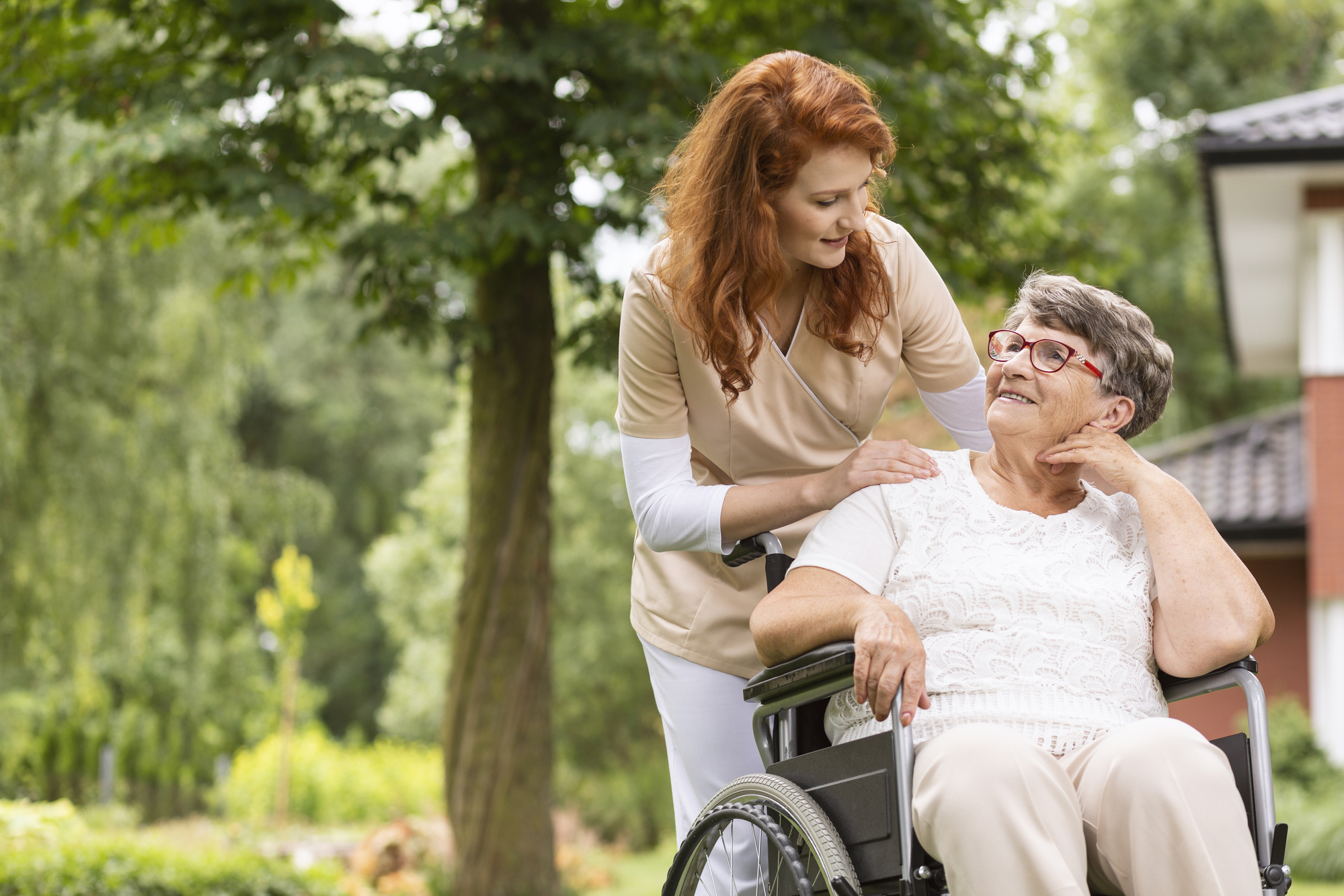 A caregiver with red hair smiles and leans over a seated elderly woman in a wheelchair. The elderly woman, wearing glasses and a white shirt, looks up at the caregiver. They are outside in a garden setting with trees and a building in the background.