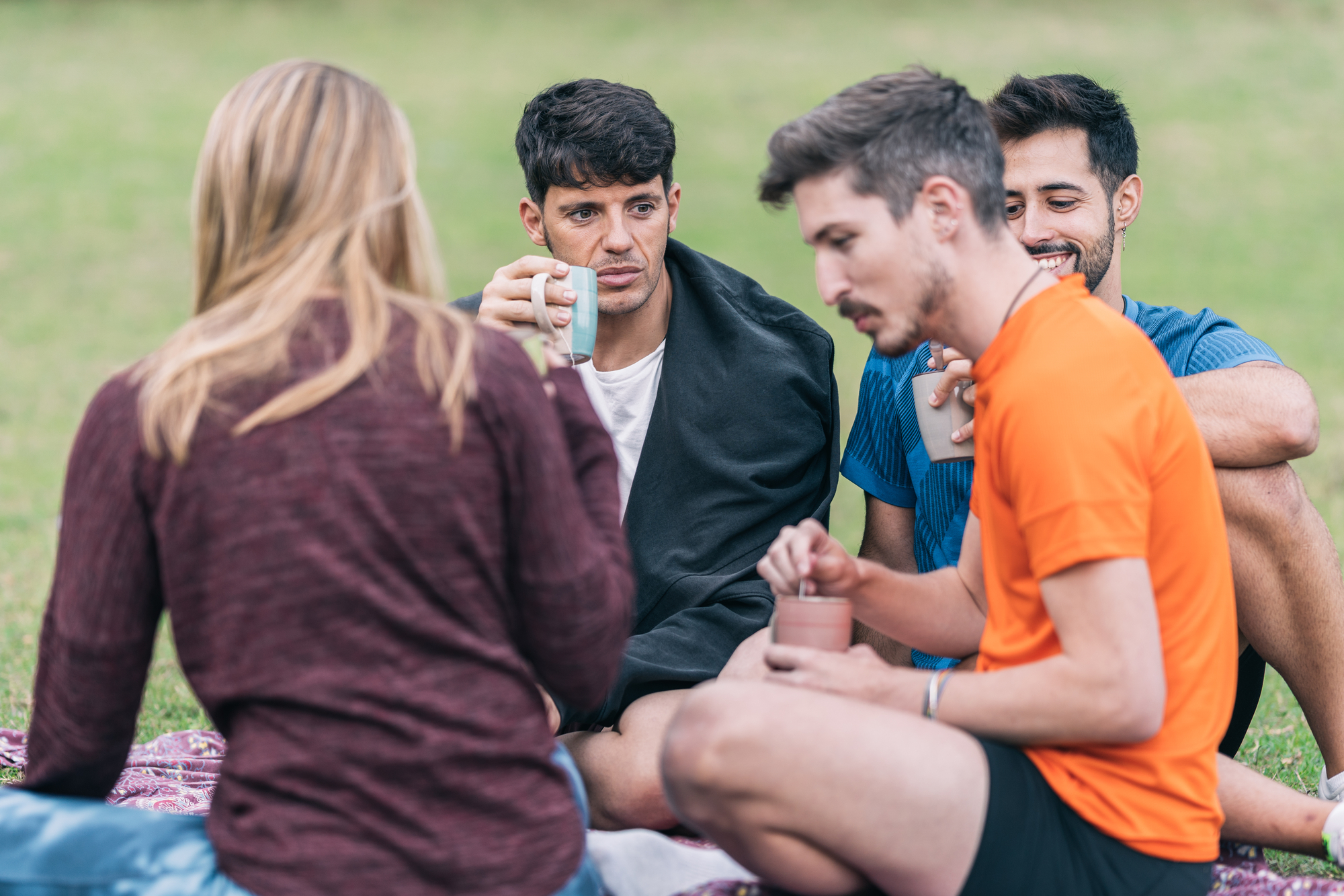 Four friends sitting on a blanket in a park, engaging in a relaxed conversation. Three men and one woman are holding cups, likely enjoying hot beverages. They are casually dressed, with the men in sportswear and the woman in a maroon sweater.