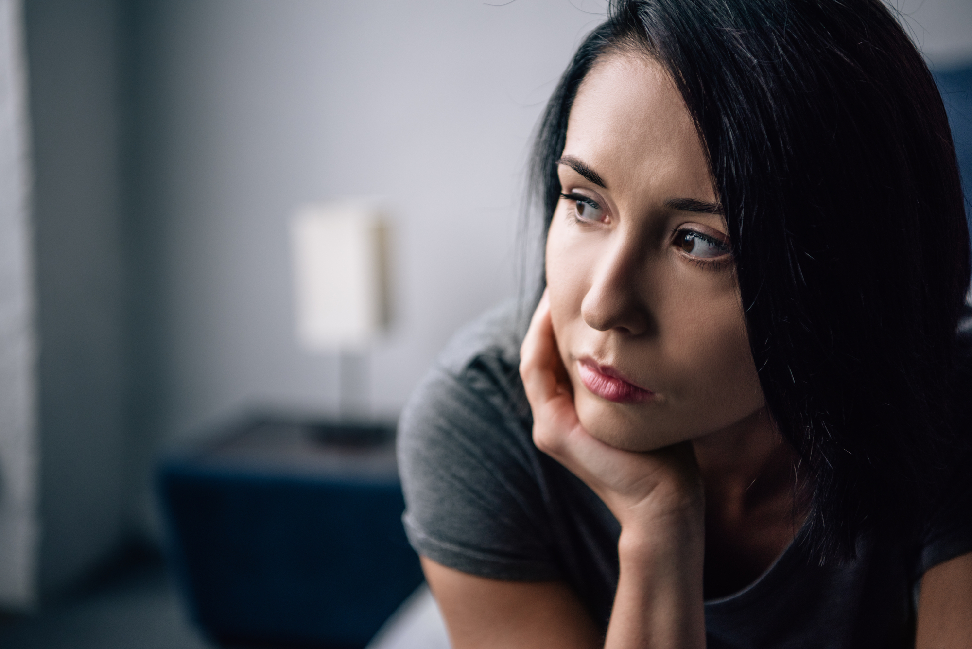 A woman with short dark hair is resting her face on her hand, looking thoughtfully into the distance. She appears pensive and is sitting indoors with a blurred background of furniture. She is wearing a gray top.