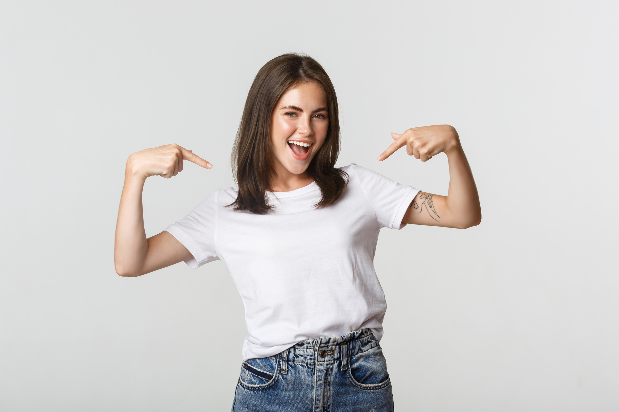 A young woman with shoulder-length brown hair stands against a plain white background. She is smiling brightly and is wearing a plain white T-shirt and blue jeans. She is pointing to herself with both hands, exuding confidence and happiness.