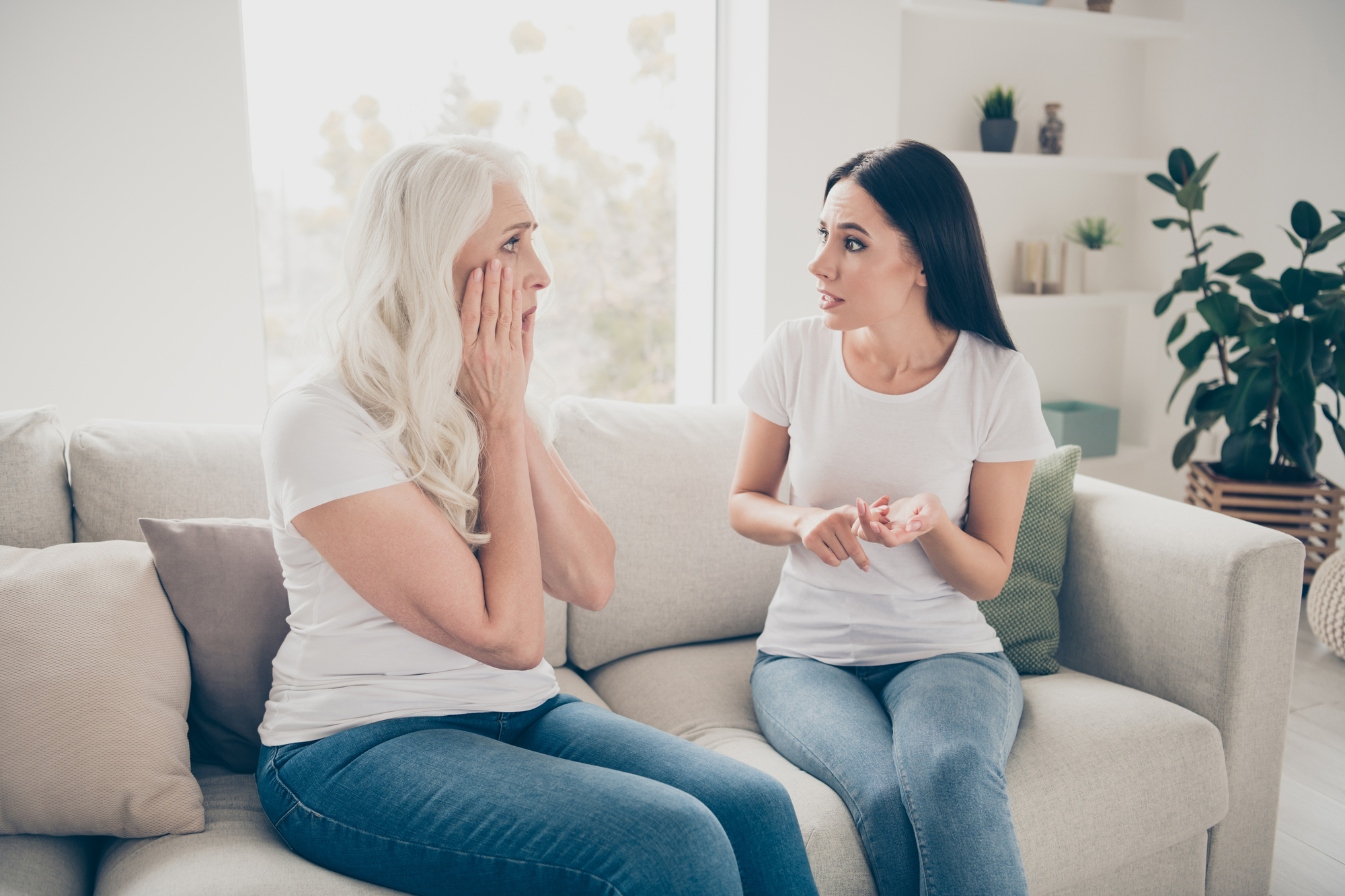 Two women are sitting on a light-colored couch in a bright room. The older woman with long white hair looks surprised, holding her face with both hands, while the younger woman with dark hair gestures expressively towards her. Both are wearing white shirts and jeans.