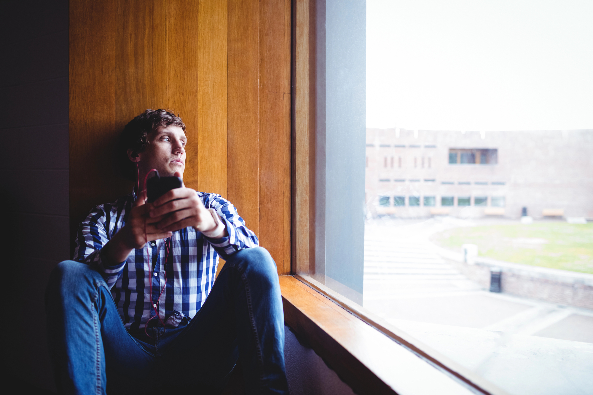 A person wearing a blue and white checkered shirt and jeans is sitting by a large window, holding a phone and looking outside with a thoughtful expression. The background shows a blurred view of buildings and an open area outside.