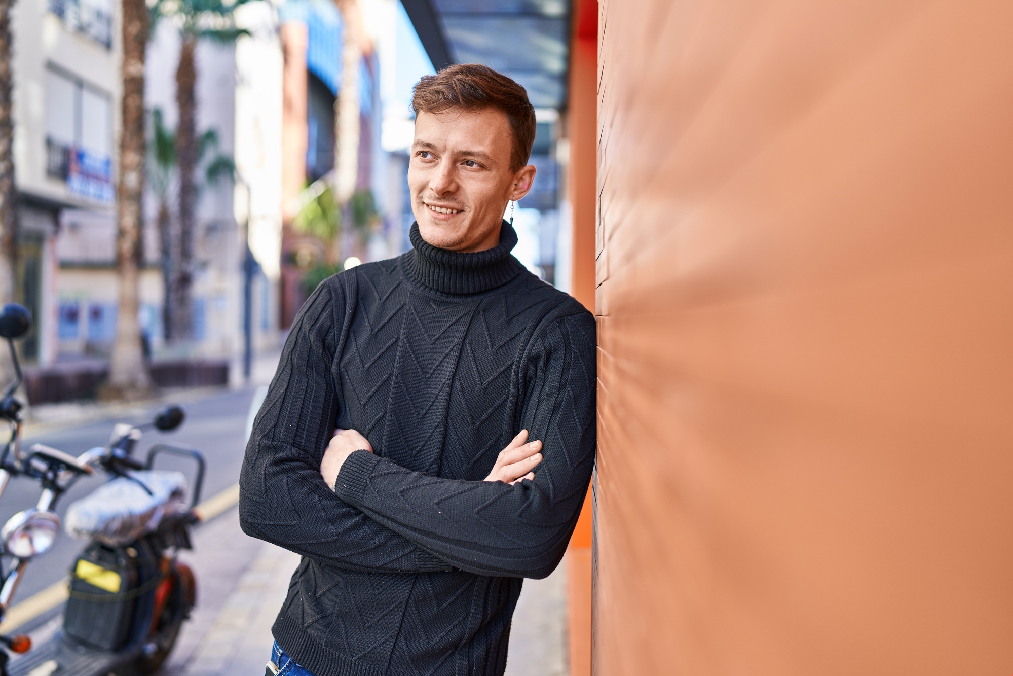 A man wearing a black turtleneck sweater stands with his arms crossed and leans against an orange wall. He is smiling and looking off into the distance. A motorbike and palm trees line the street in the background. The scene is sunny and urban.