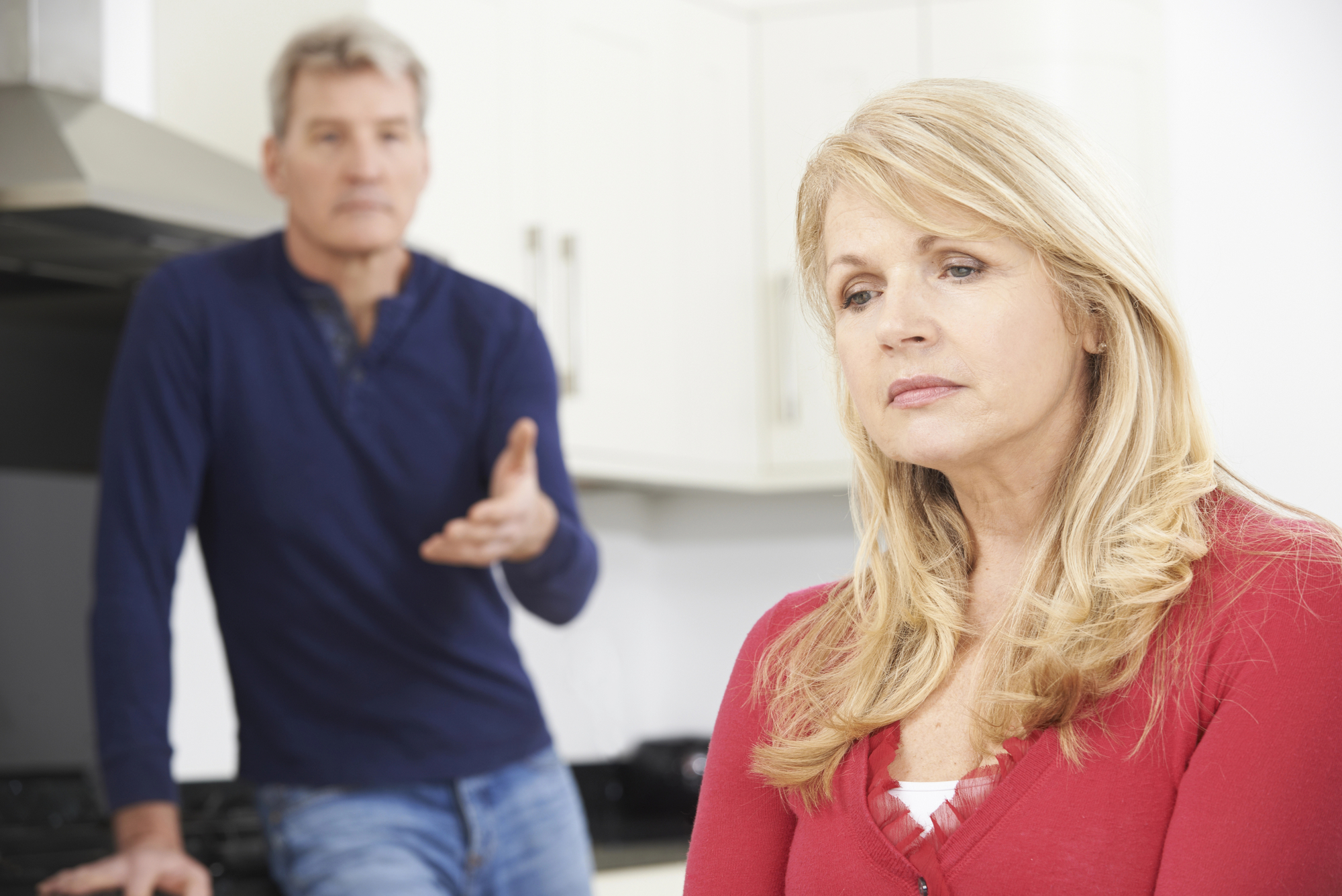 A woman with long blonde hair, wearing a red sweater, stands looking pensive in a kitchen. A man in the background is slightly out of focus with gray hair, wearing a blue shirt, appears to be talking, gesturing with one hand.