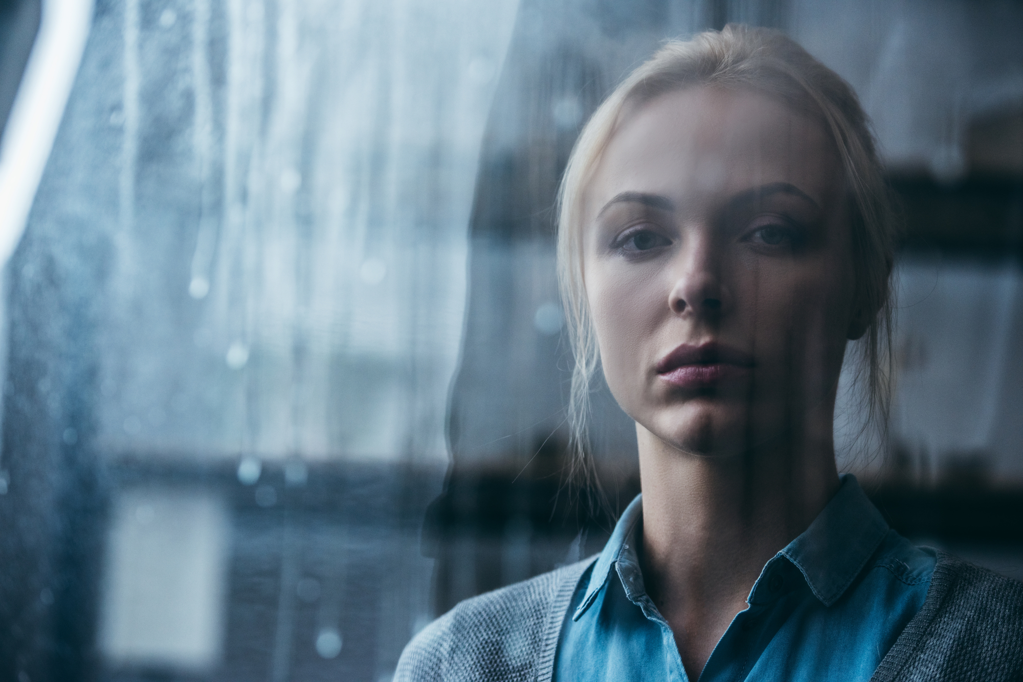 A woman with blonde hair is standing behind a rain-streaked window. She is wearing a blue shirt and a gray cardigan, looking pensively into the distance. The background is blurred and out of focus.