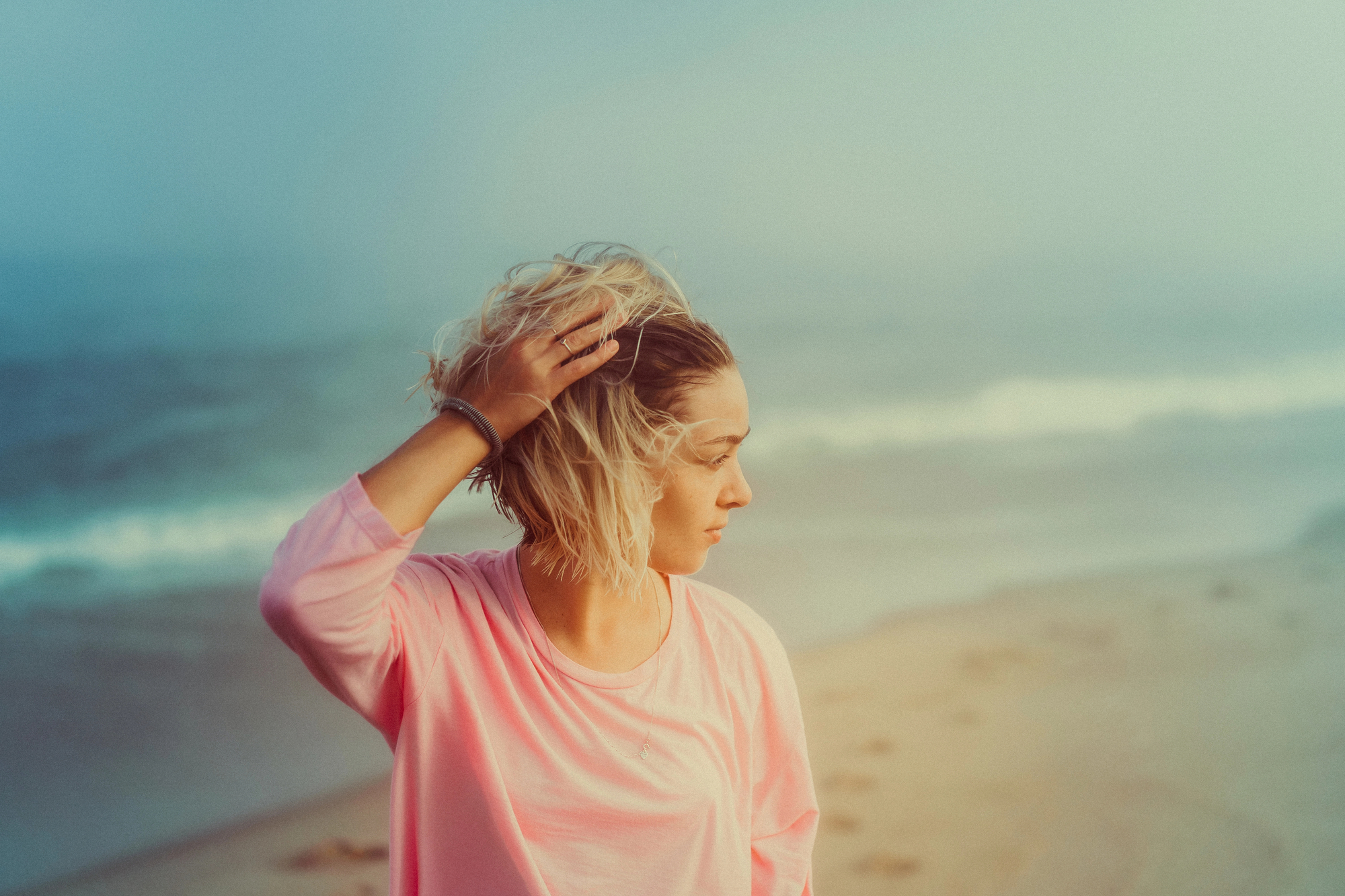 A woman with blonde hair, wearing a pink long-sleeved top, stands on a beach with her hand in her hair. She looks to the side with the ocean and waves in the background, and footprints are visible in the sand. The sky is hazy, creating a serene atmosphere.