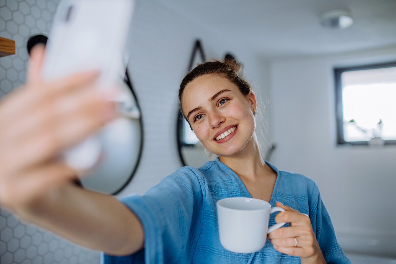 A young woman wearing a blue top stands indoors holding a white mug in one hand and taking a selfie with the other. She smiles at her phone's camera. The background shows a white tiled wall and a small window.