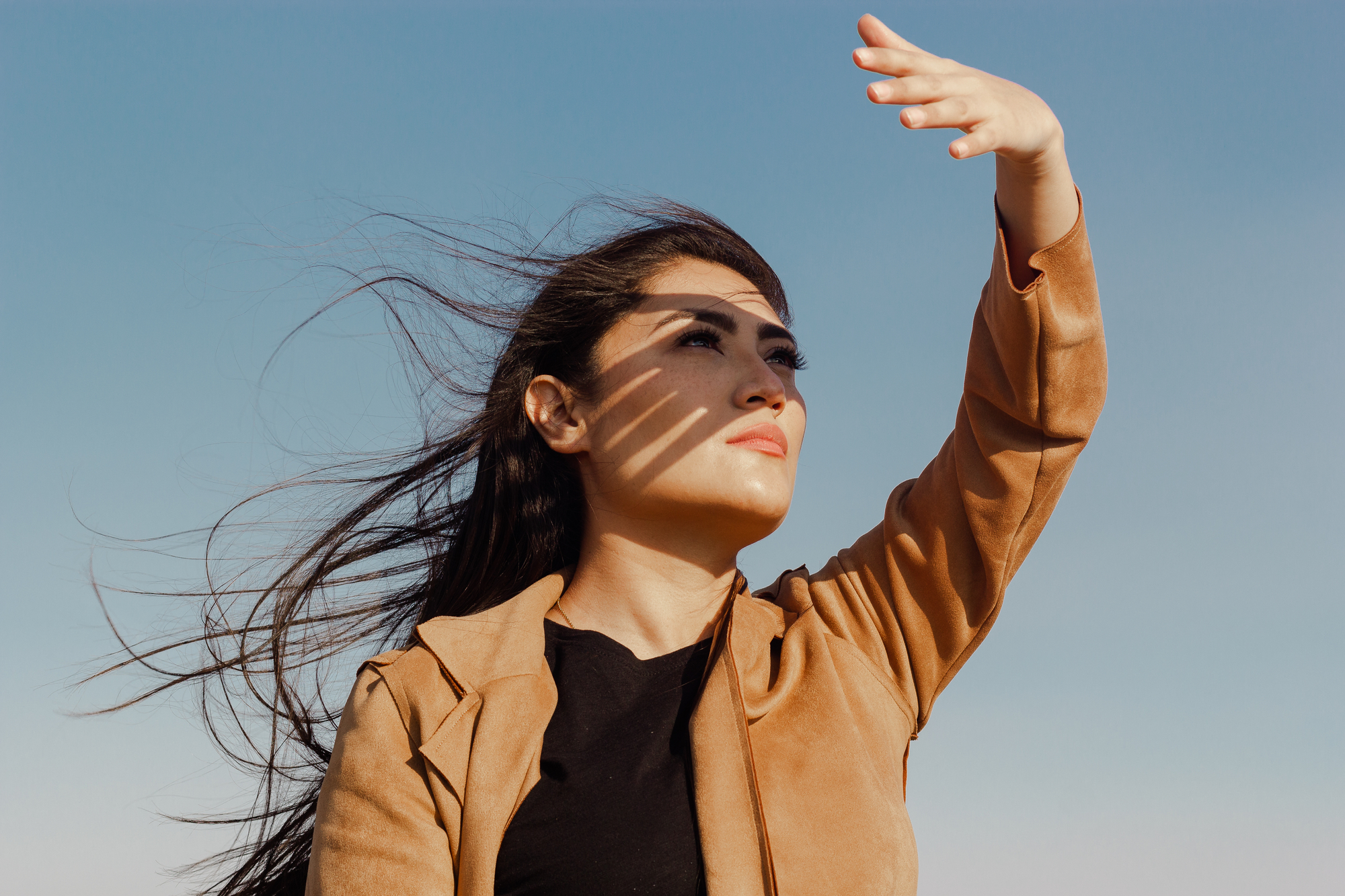 A woman with long hair, wearing a brown jacket and black shirt, raises her hand to shield her eyes from the sun. Shadow patterns cross her face as she looks upwards with a serene expression. The blue sky in the background emphasizes the bright, sunny day.