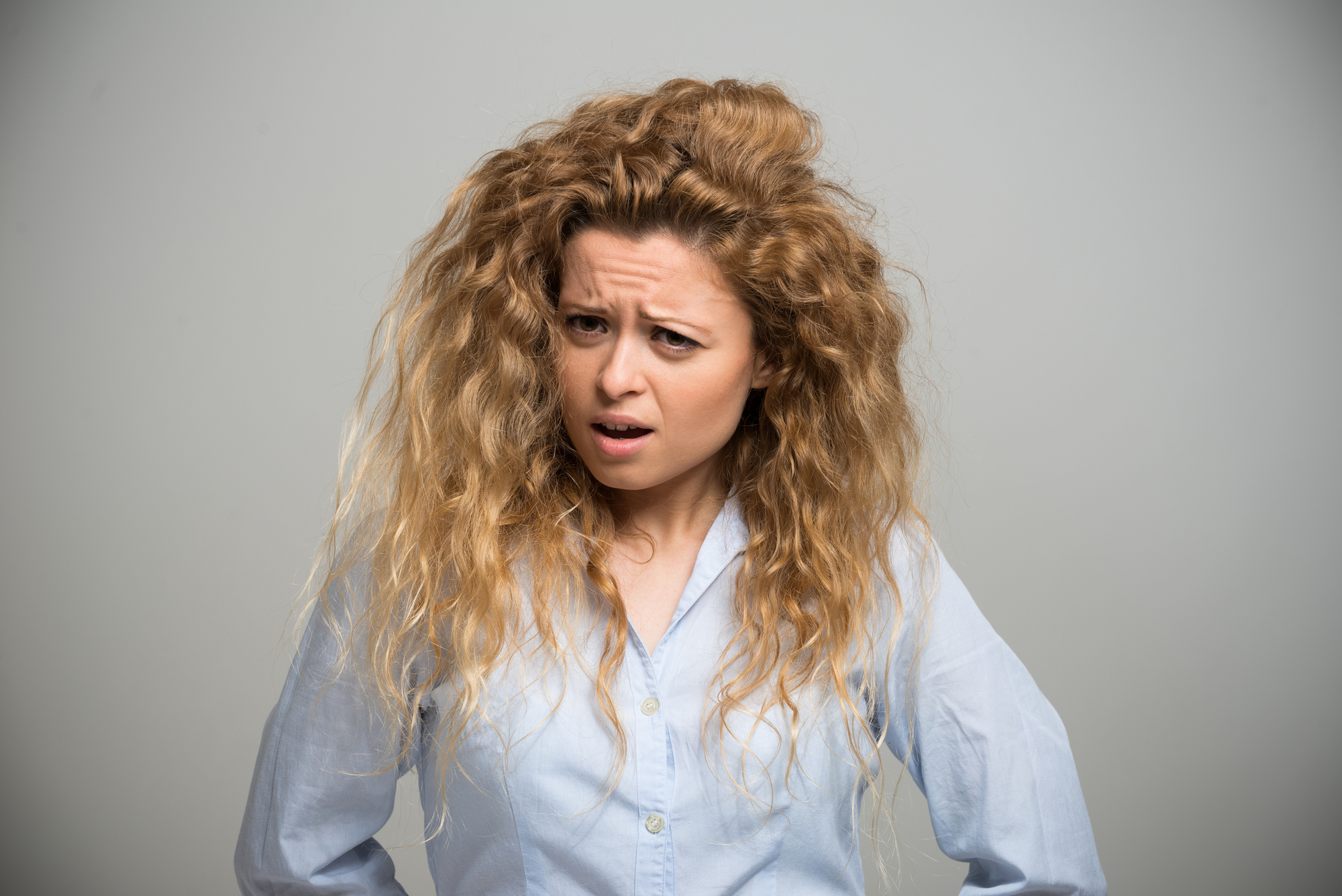 A woman with long, curly hair and a light blue shirt stands against a light gray background. She has a slightly furrowed brow and a puzzled or questioning expression.