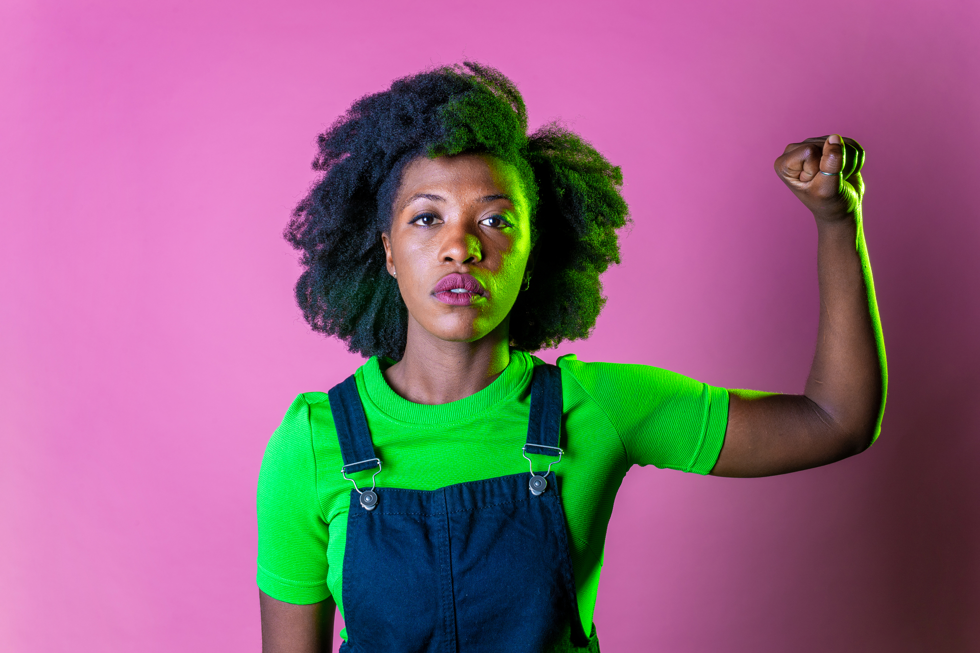 A person with curly hair, wearing a bright green shirt and denim overalls, stands against a pink background. They raise their right arm with a clenched fist in a gesture of strength and determination.