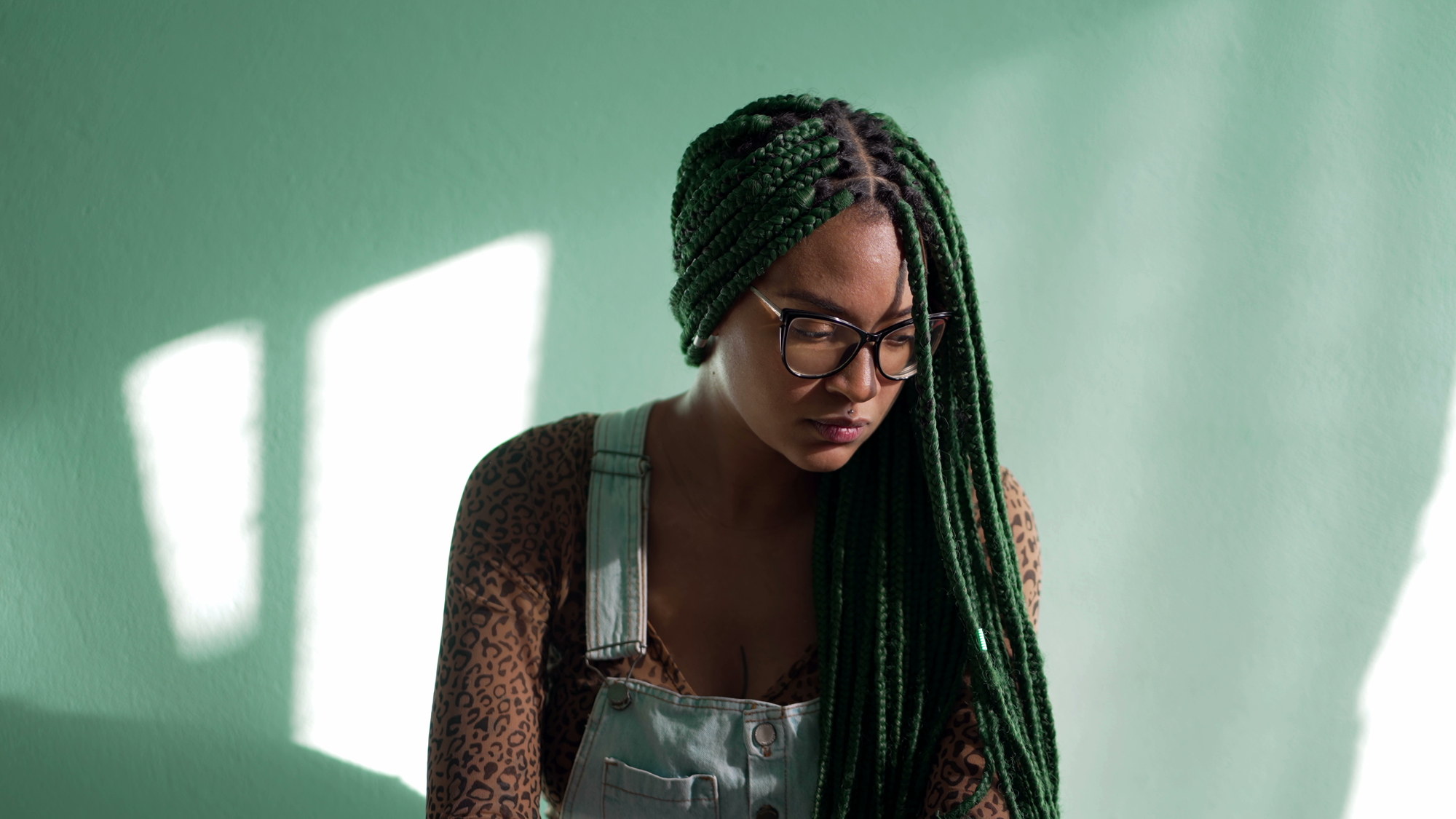 A woman with long, green braids and glasses sits against a green wall. She is wearing a patterned top and denim overalls. The lighting casts a bright shadow on the wall, highlighting her contemplative expression.