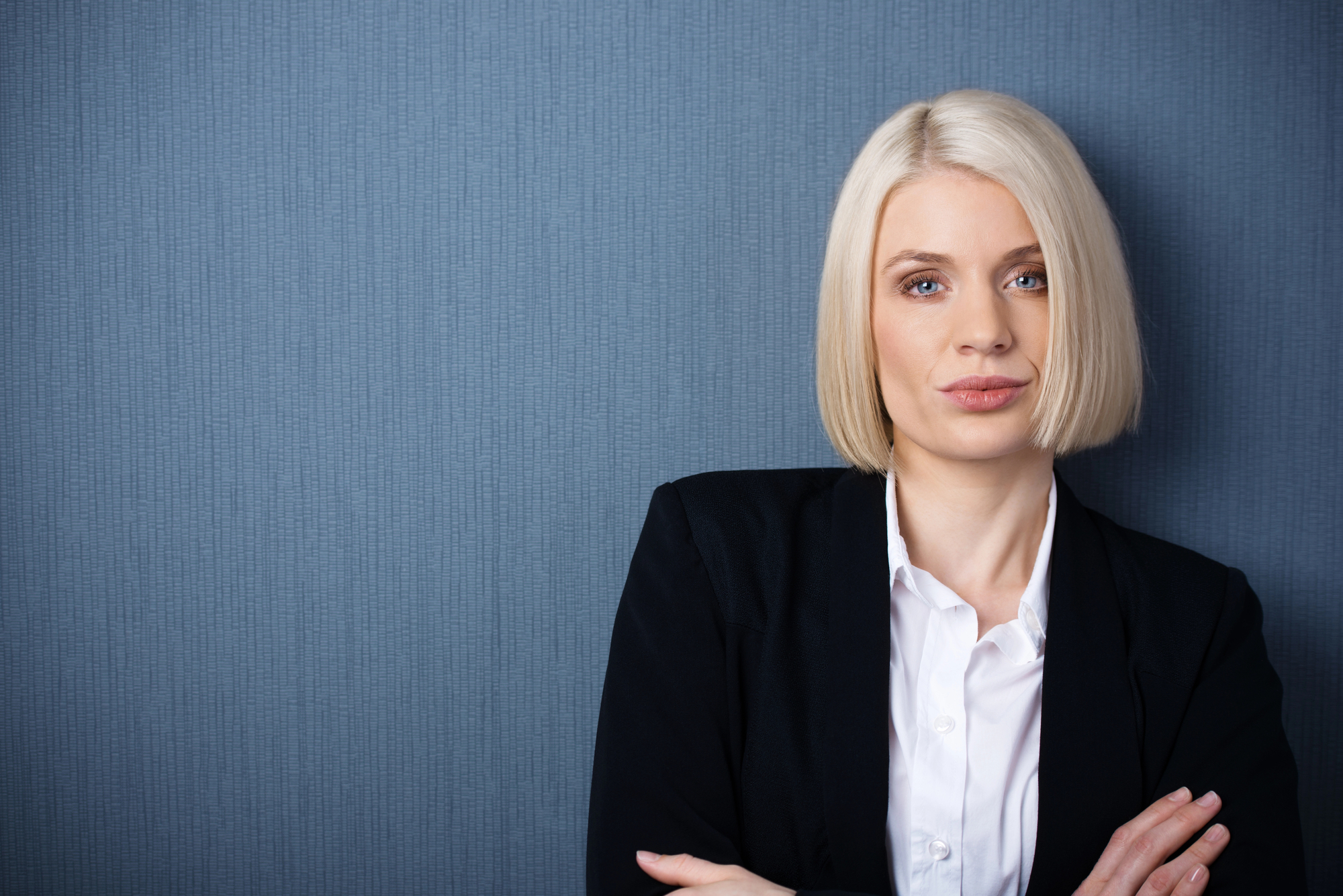 A woman with straight blonde hair cut into a bob stands against a textured blue wall. She is wearing a white collared shirt and a black blazer, with her arms crossed, gazing directly at the camera with a neutral expression.