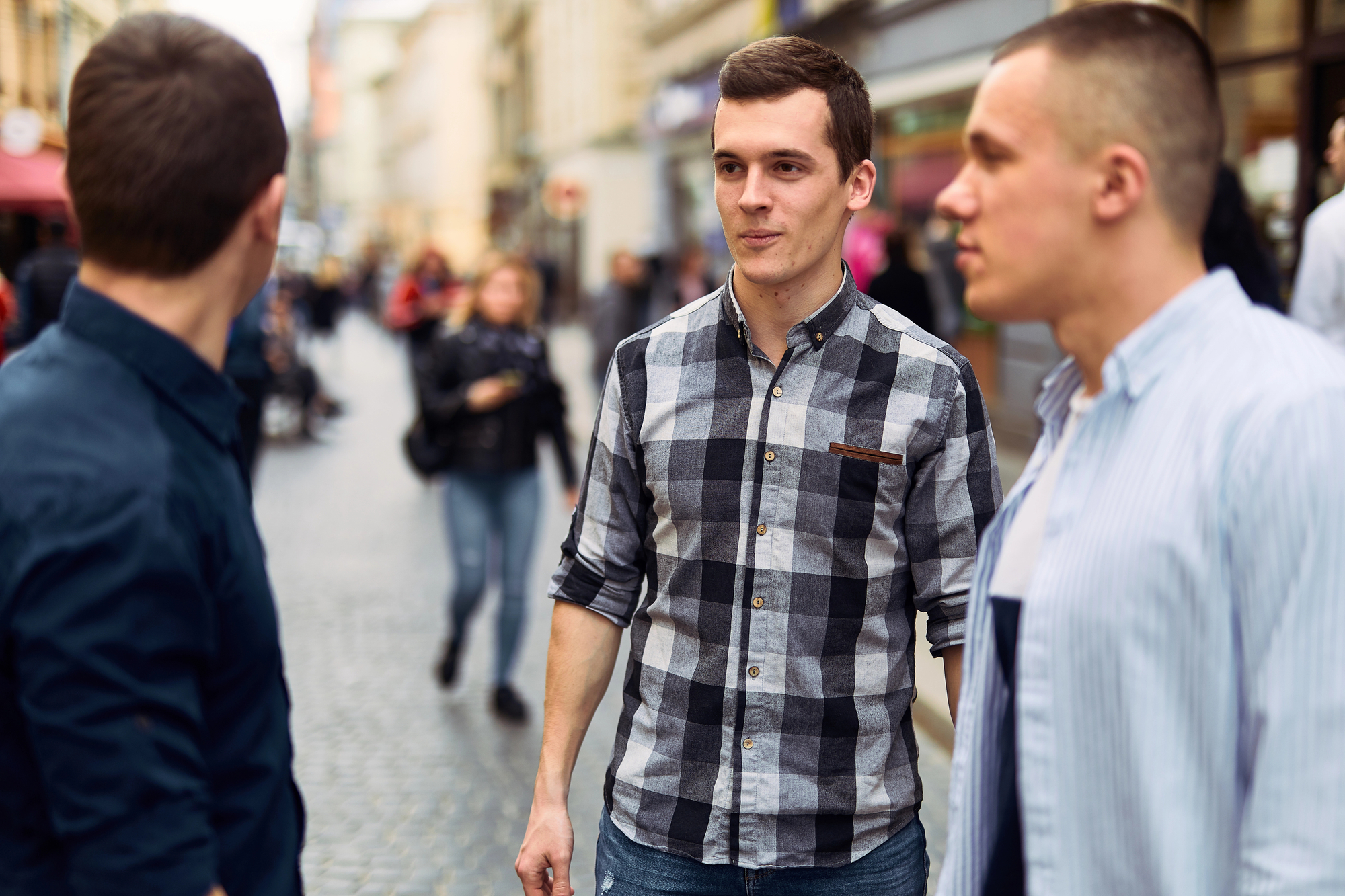 Three young men are standing and talking on a city street with various people walking in the background. The man in the middle is wearing a gray checked shirt, the one on the left is in a dark blue shirt, and the person on the right is in a light blue shirt.