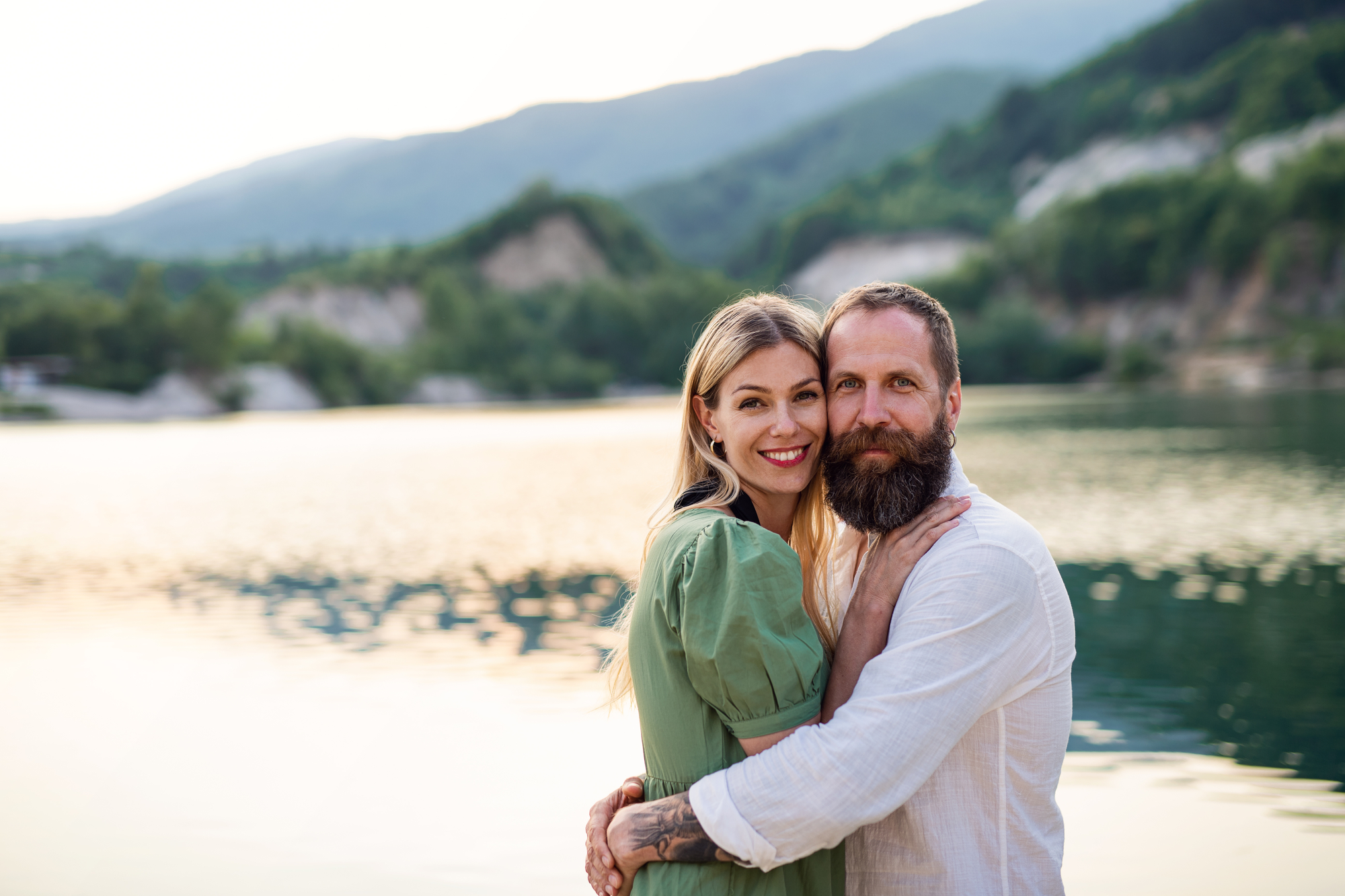 A couple stands by a serene lake with mountains in the background. The woman, wearing a green dress, smiles and rests her hand on the chest of the man with a beard and white shirt. Both look at the camera, enjoying the peaceful, sunset-lit scene.