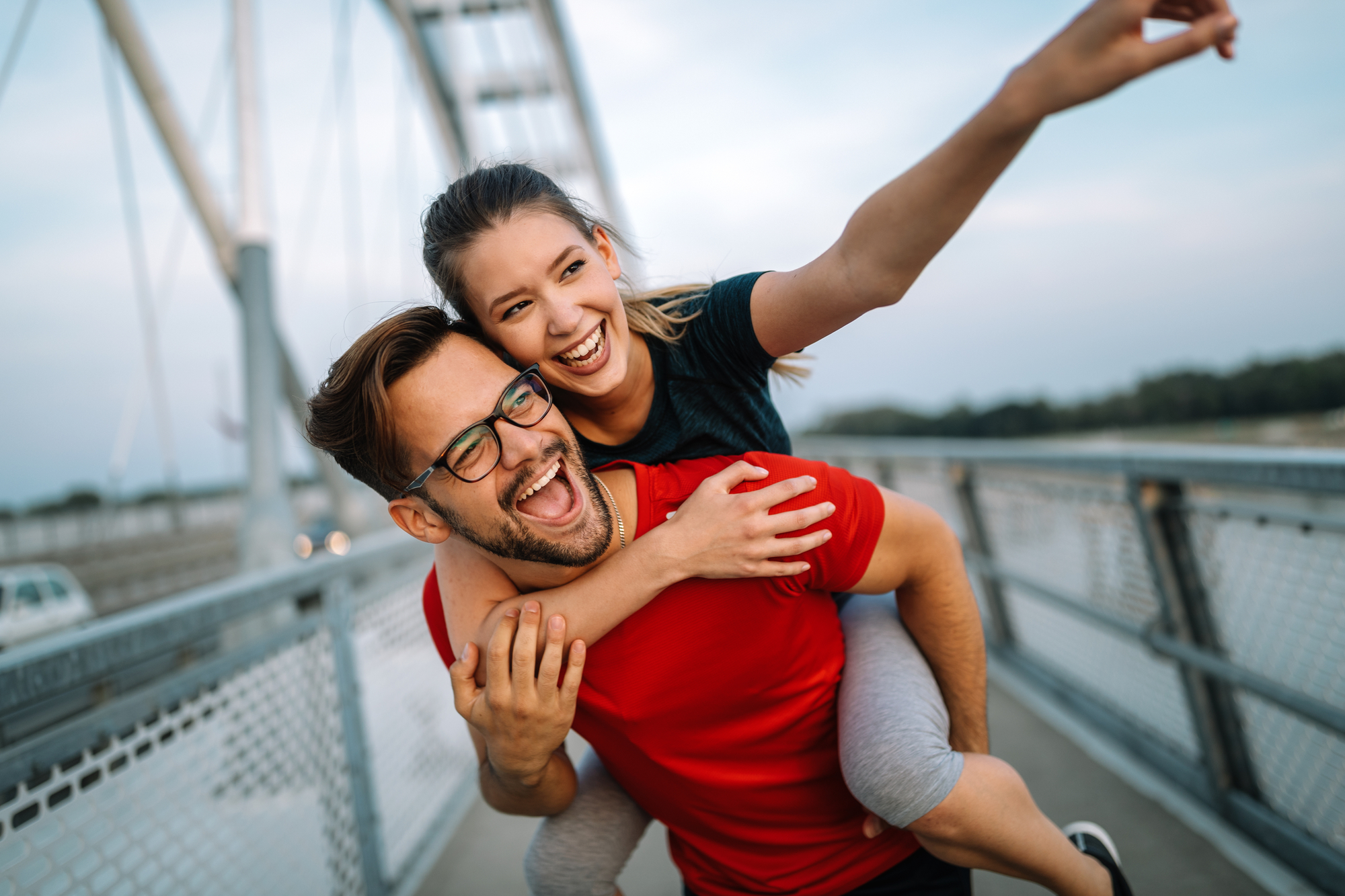 A joyful couple enjoys a playful moment on a bridge. The woman, dressed in athletic wear, is piggybacking on the man, who is wearing a red shirt and glasses. She points excitedly into the distance, while both of them have wide, happy smiles. The background shows the bridge structure.