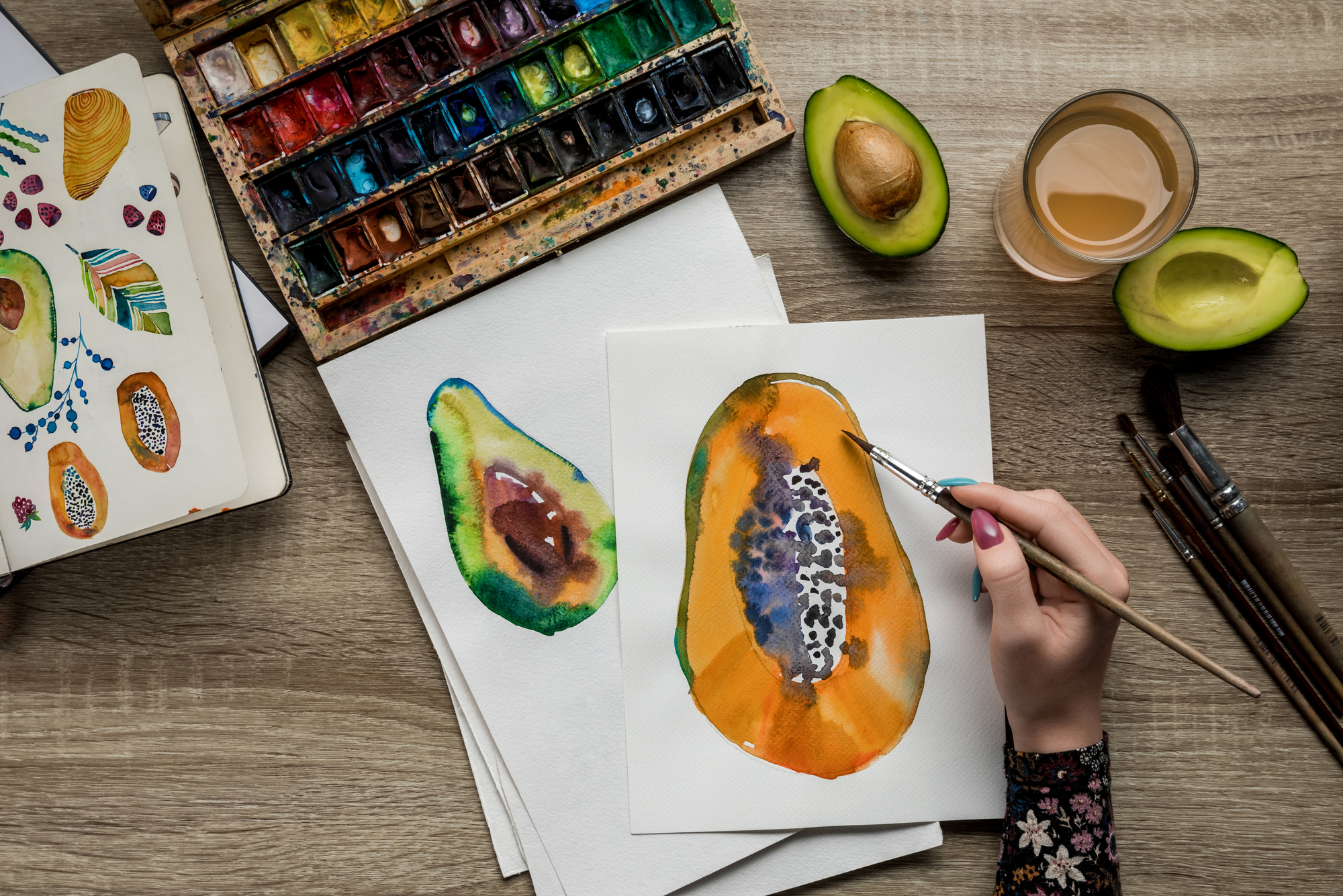 A person painting a watercolor image of a papaya on white paper. The table has watercolors, paintbrushes, a glass of juice, and avocado halves. Another painting of a papaya on the left serves as a reference.