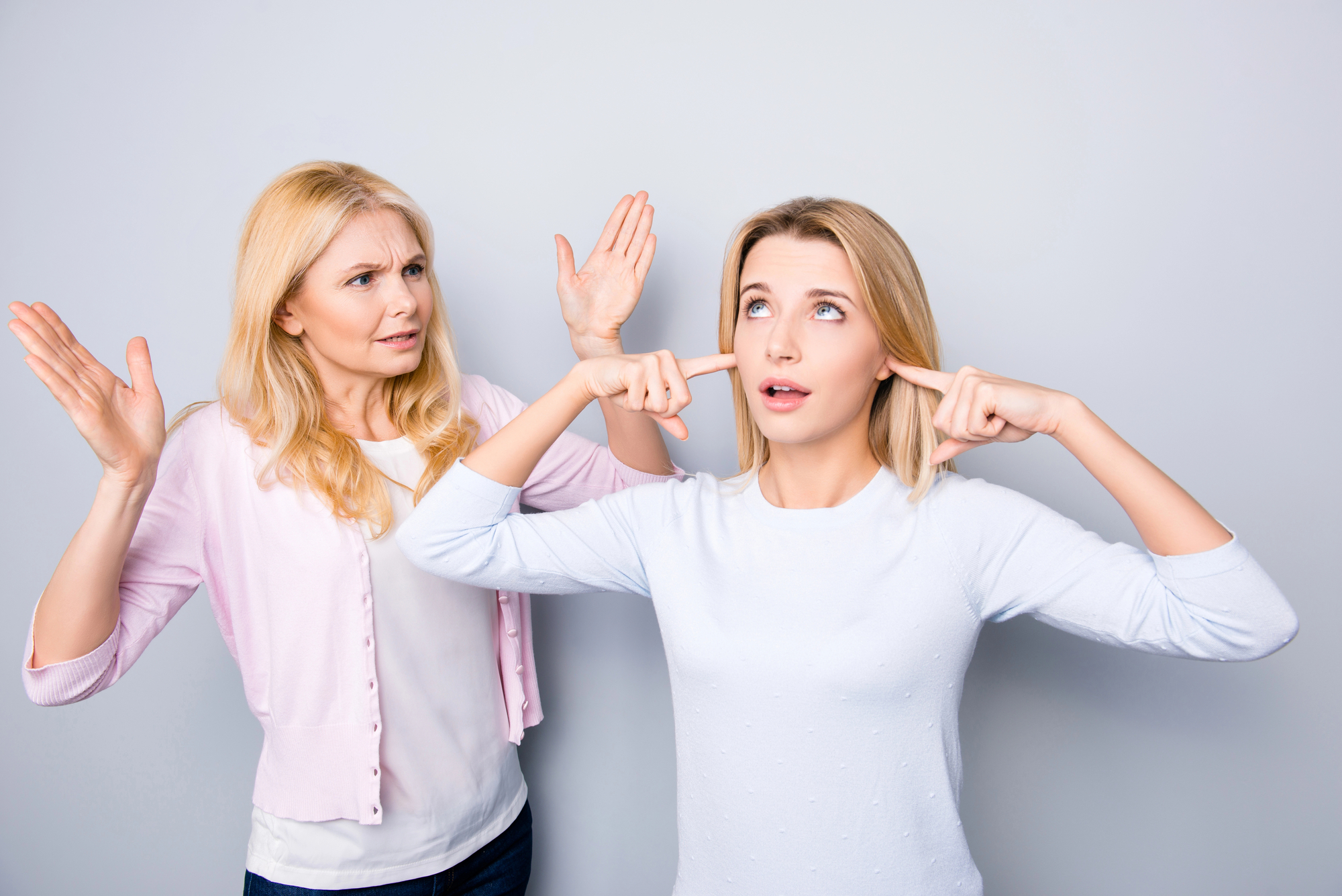 A woman on the left is angrily gesturing at a younger woman on the right, who is ignoring her by plugging her ears with her fingers and looking upwards. Both have long blonde hair and are wearing casual clothes against a plain background.