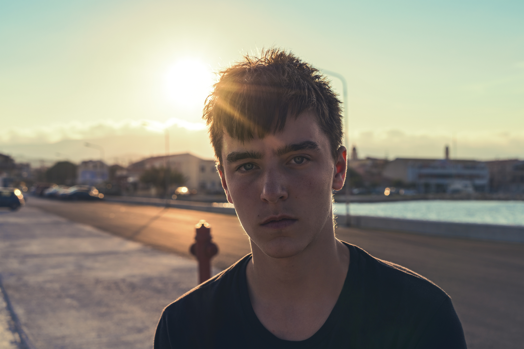 A young person with short, dark hair is standing on an outdoor street at sunset. The sun is low in the sky, casting a warm glow and lens flare. Buildings and a body of water are visible in the background. The person is wearing a dark shirt and has a serious expression.