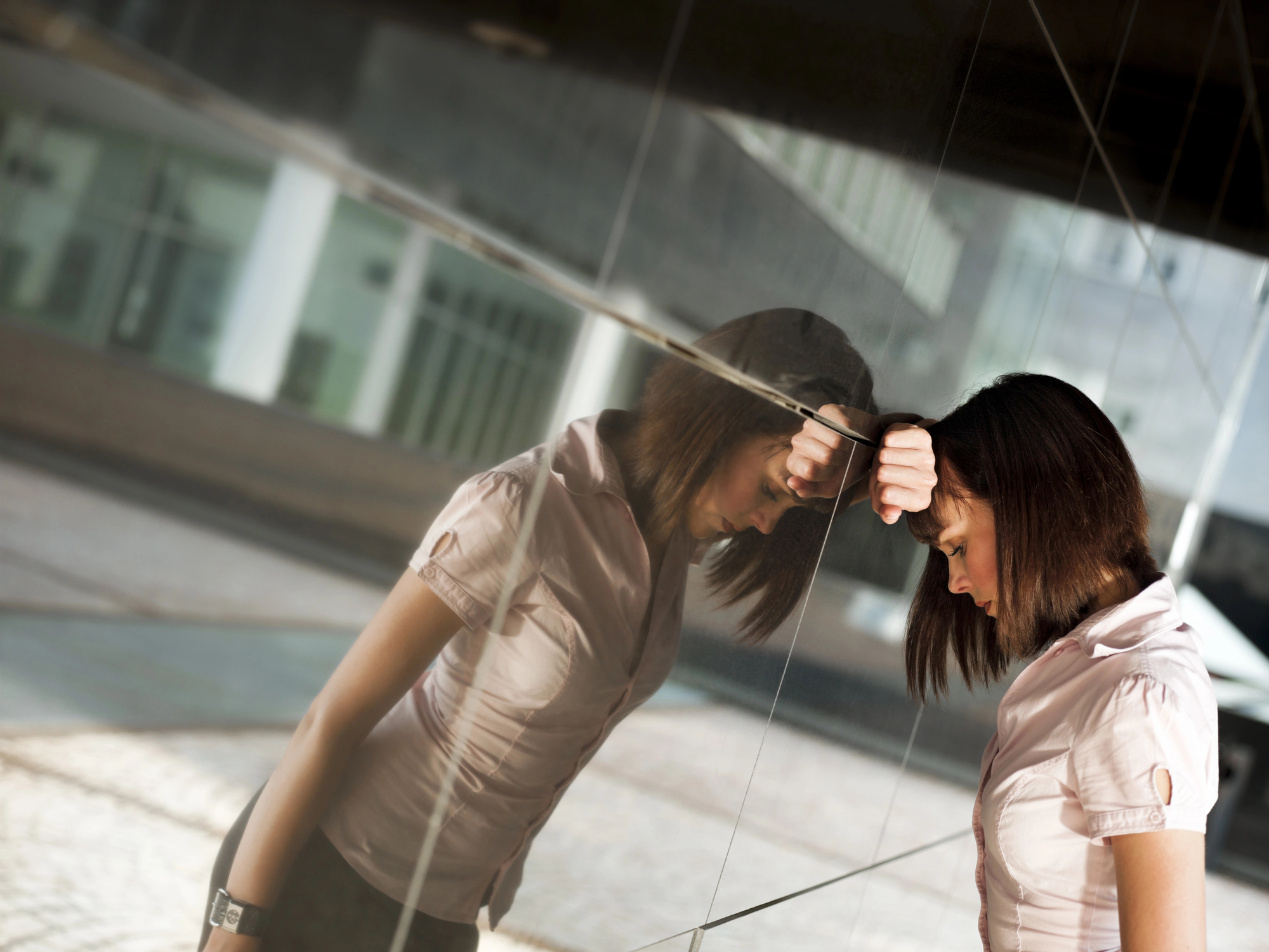A woman in a short-sleeve blouse rests her forehead against a reflective glass wall, appearing contemplative or distressed. Her reflection is visible in the glass, and the background shows an urban setting with buildings and a paved surface.