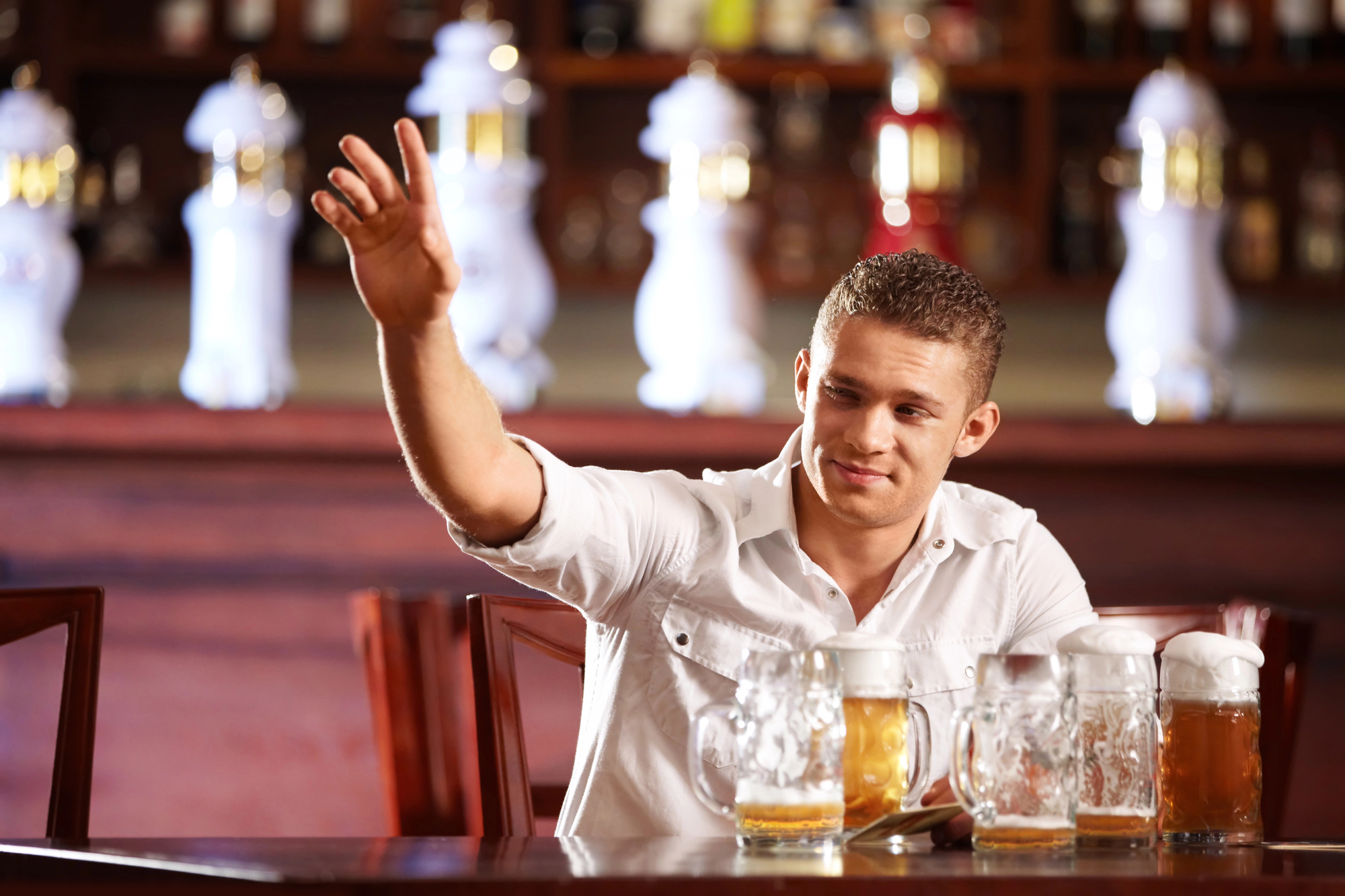 A man in a white shirt is sitting in a bar with several glass mugs of beer on the table in front of him. He is smiling, looking to his left, and raising his right hand as if waving or signaling. The background features a bar with various bottles.