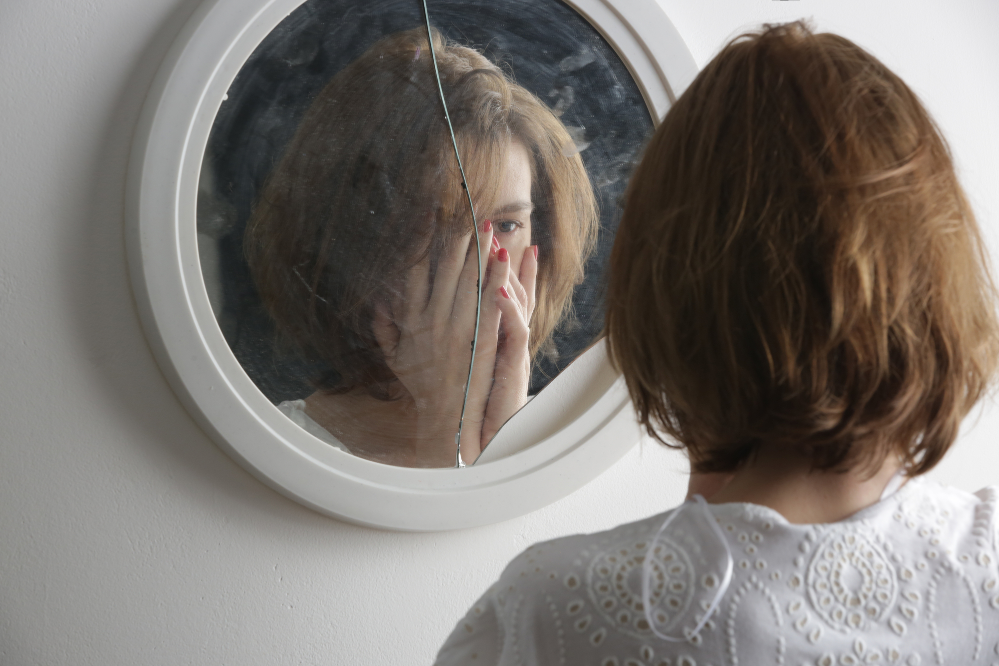 A person with short, brown hair is standing in front of a cracked round mirror, looking at their reflection. They are wearing a white shirt with intricate patterns, and their hand is partially covering their face. The background is a plain white wall.