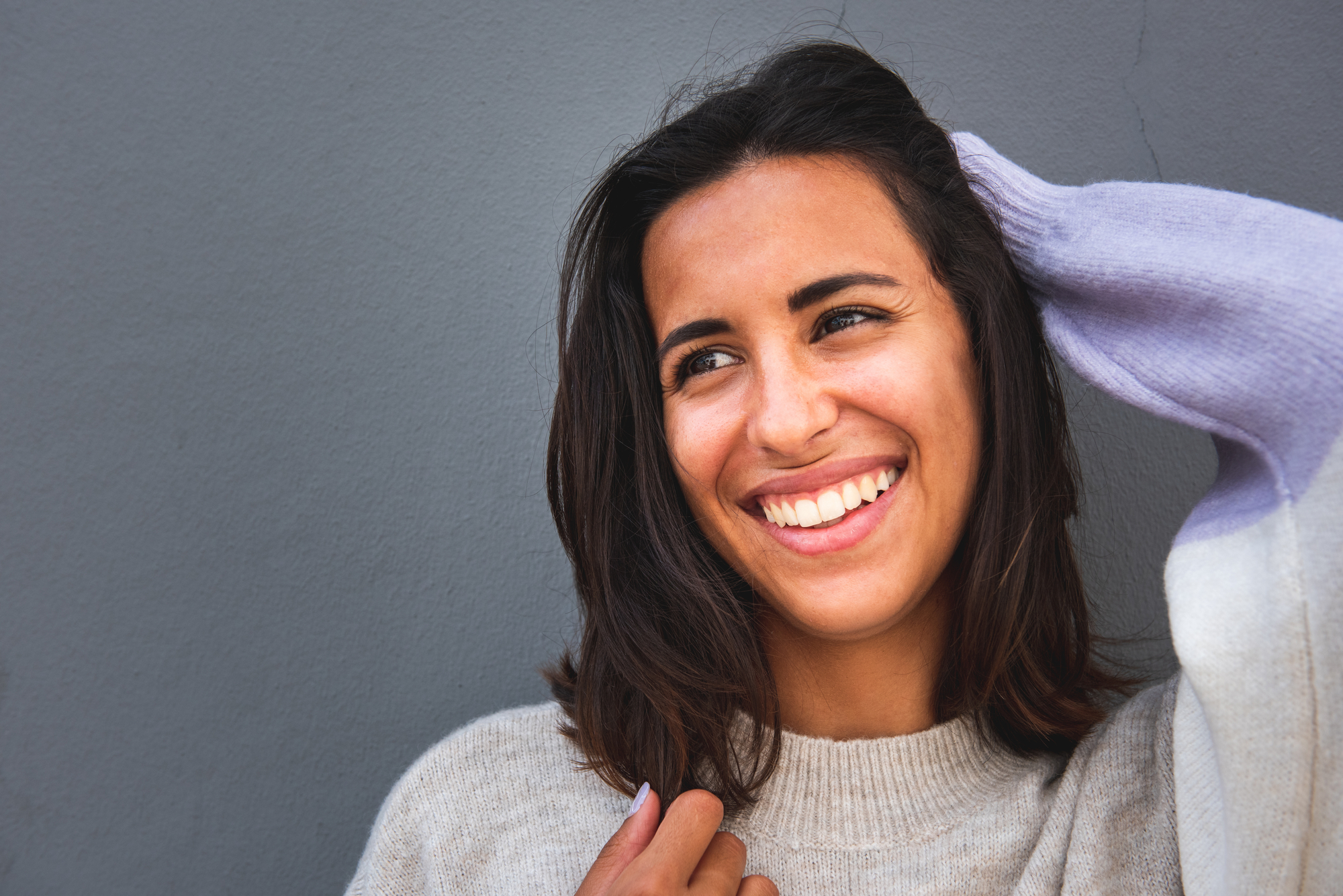 A young woman with shoulder-length dark hair smiles warmly while holding one hand up to her head. She is wearing a light-colored sweater and stands against a plain gray background.
