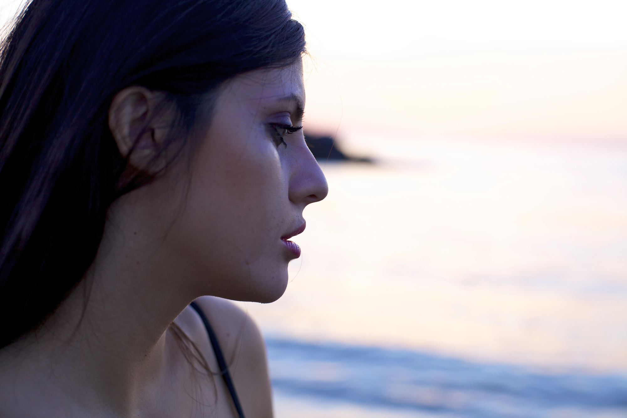 A profile view of a woman with long dark hair looking contemplatively towards the sea at sunset. The calm ocean and gentle colors of the sunset create a serene and introspective atmosphere.