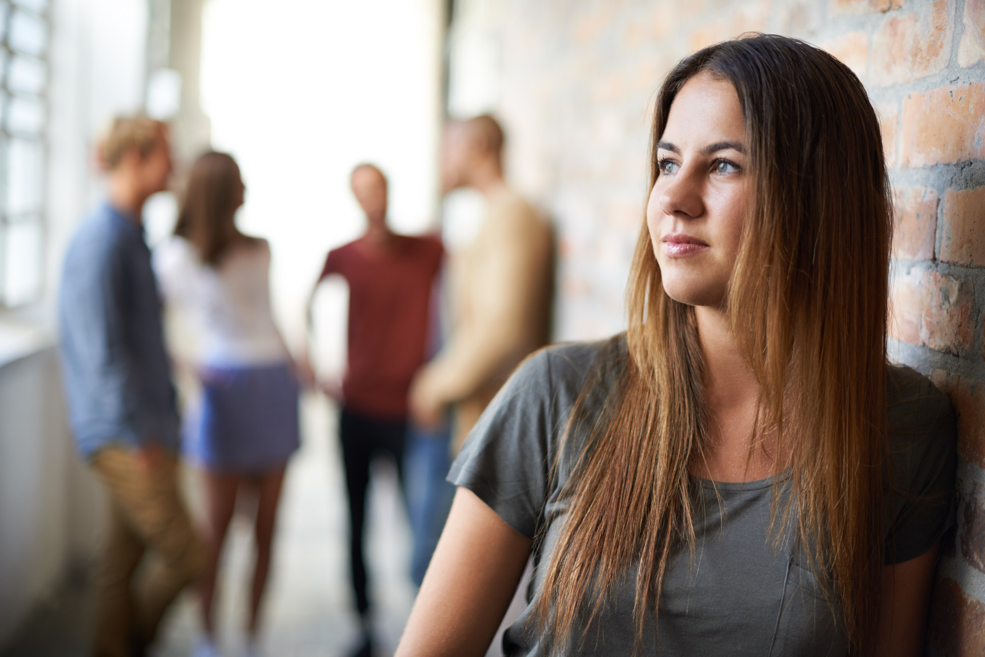 A young woman with long brown hair leans against a brick wall, looking pensively into the distance. In the background, a group of four people is engaged in conversation, with their faces blurred and the focus on the woman in the foreground.