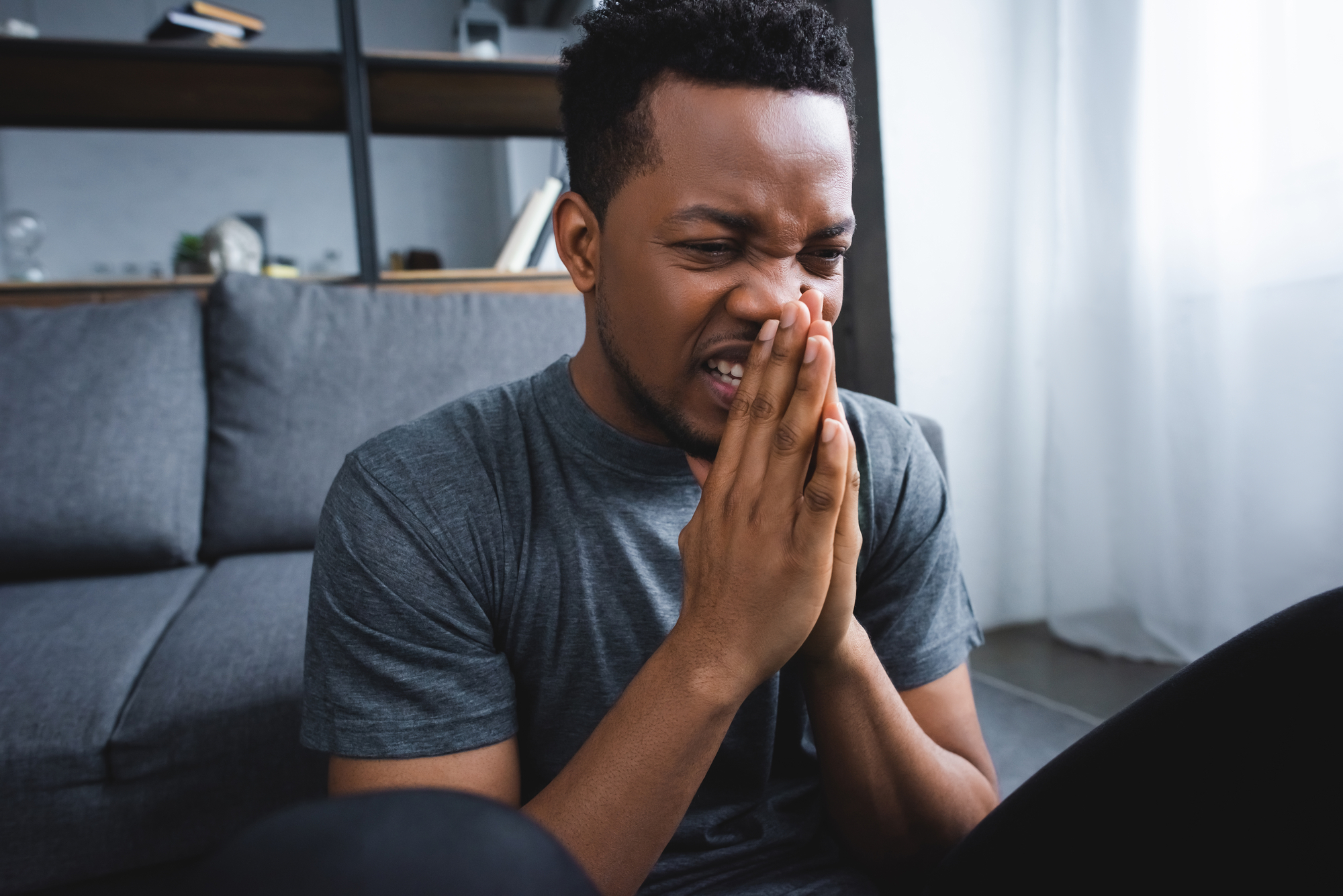 A person in a gray t-shirt sits on the floor with hands clasped in front of their face, appearing to be in deep thought or distress. There's a gray sofa and a wooden shelf in the background, next to a sunlit window with white curtains.