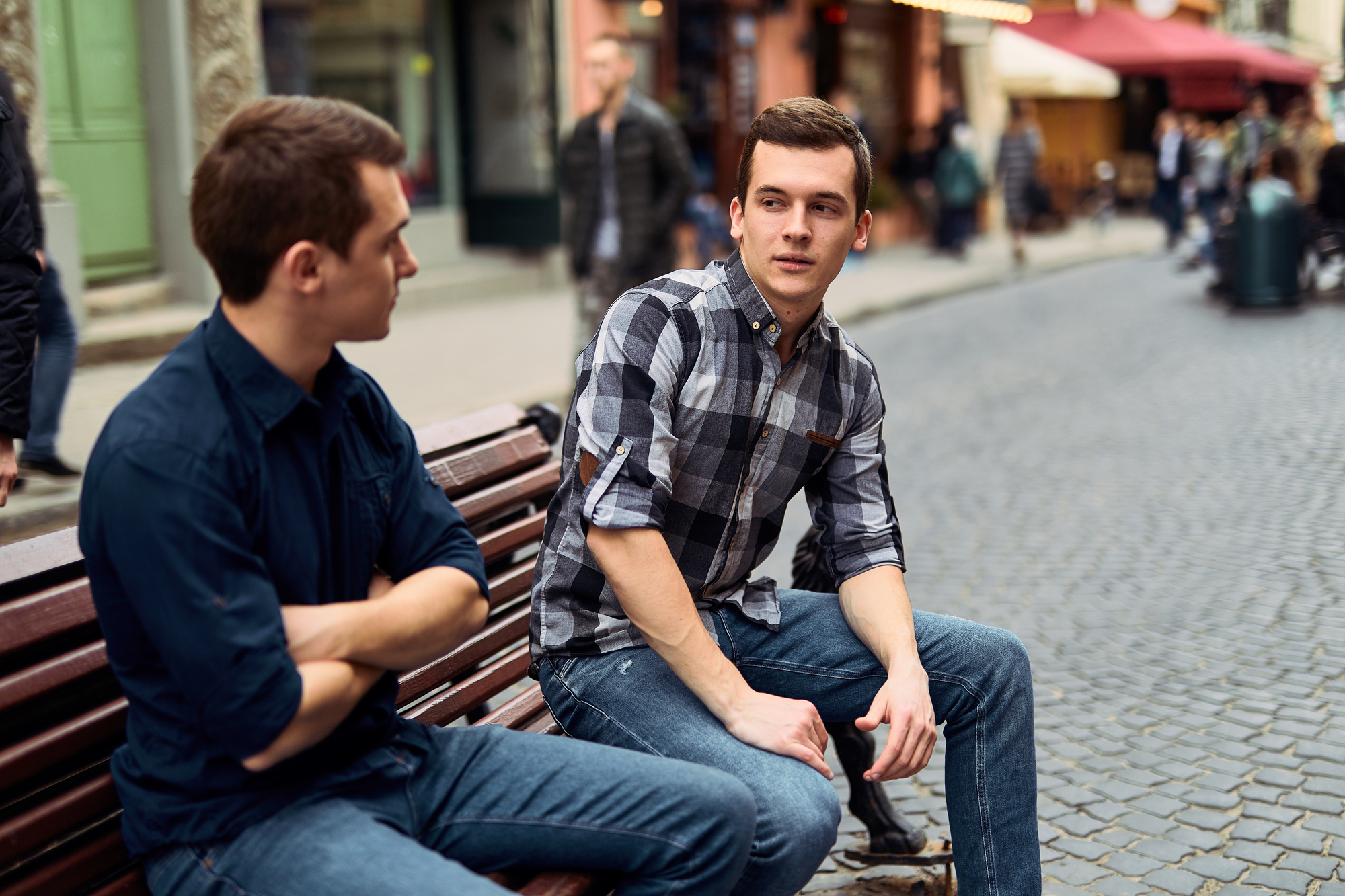 Two young men are sitting on a wooden bench on a cobblestone street. One is wearing a navy blue shirt with his arms crossed and facing the other, who is in a grey plaid shirt, engaged in conversation. Both have short hair and are dressed casually in jeans.