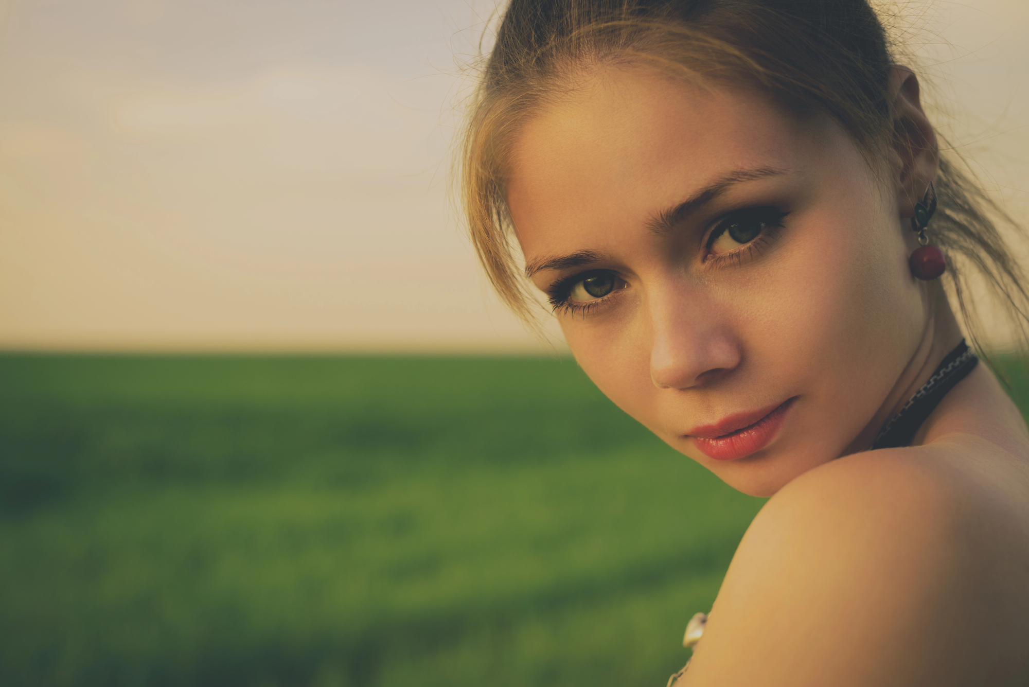 A young woman with light brown hair pulled back gazes softly towards the camera. She is outdoors with a blurred background of a green field and a soft, cloudy sky. She wears subtle makeup and earrings, with one shoulder exposed in the warm light.