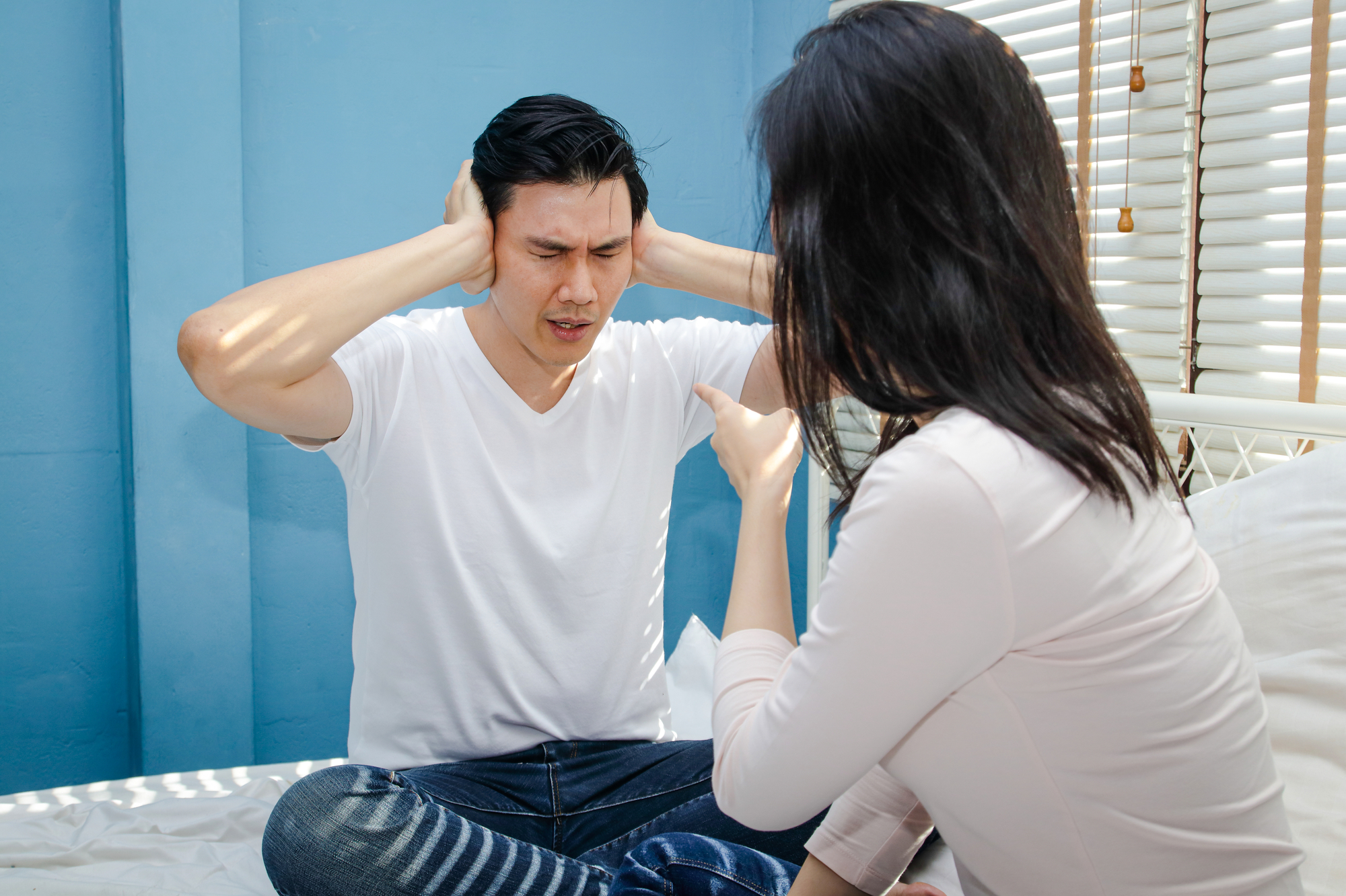 A man sitting on a bed with a distressed expression, covering his ears with his hands. A woman, also sitting on the bed and facing him, is pointing and appears to be speaking or yelling at him. The setting is a room with blue walls and a window with blinds.