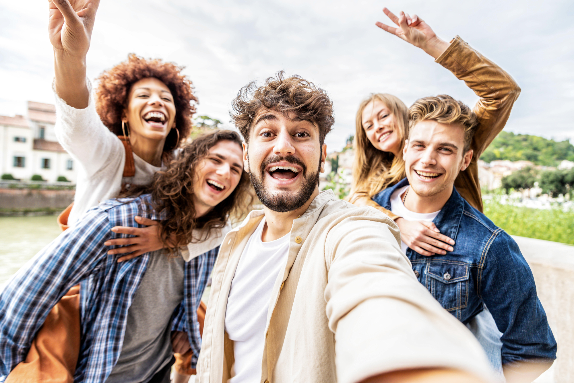A group of five friends taking a selfie outdoors. They are all smiling and appear joyful. One person is giving a piggyback ride, while another is making a peace sign with their hand. They are standing against a backdrop of buildings and greenery.