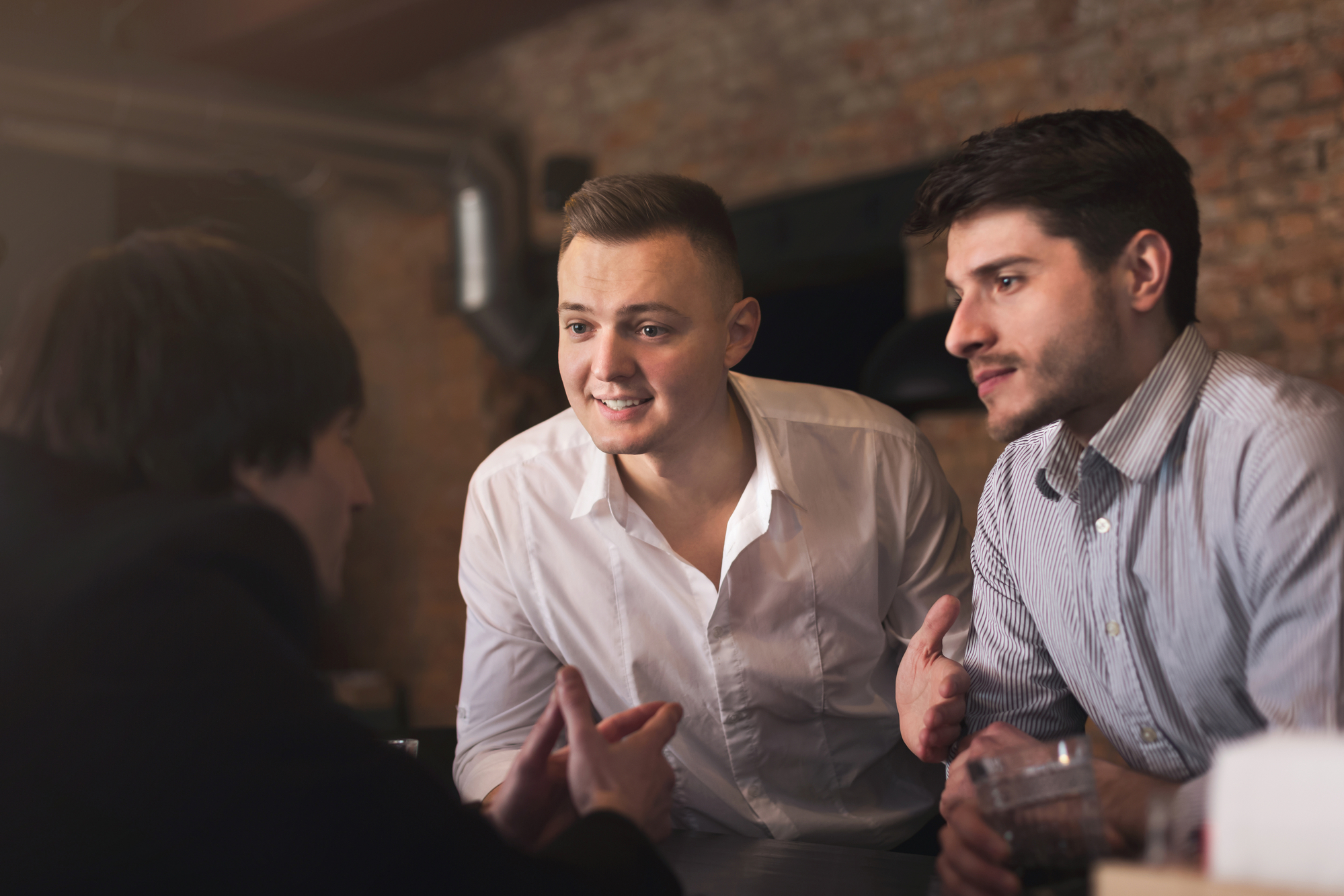 Three men are engaged in a conversation in a dimly lit setting with an exposed brick wall in the background. Two men in white and patterned button-down shirts are facing a man with dark hair, who is seen from behind. They appear animated and focused.