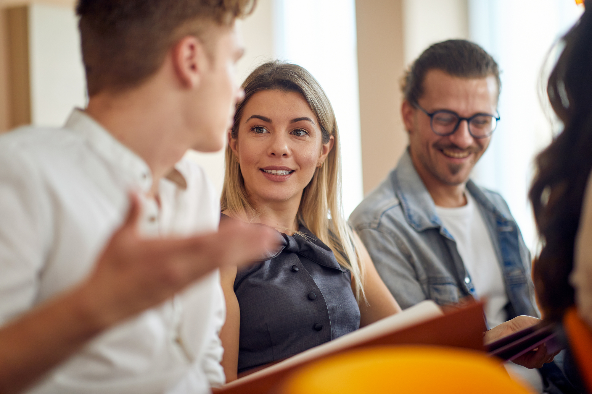 Three people are seated and engaged in conversation. A woman in the center, wearing a gray dress, smiles while listening. Two men, one on each side of her, are also part of the discussion. The man on the left is gesturing with his hand, while the man on the right is smiling.