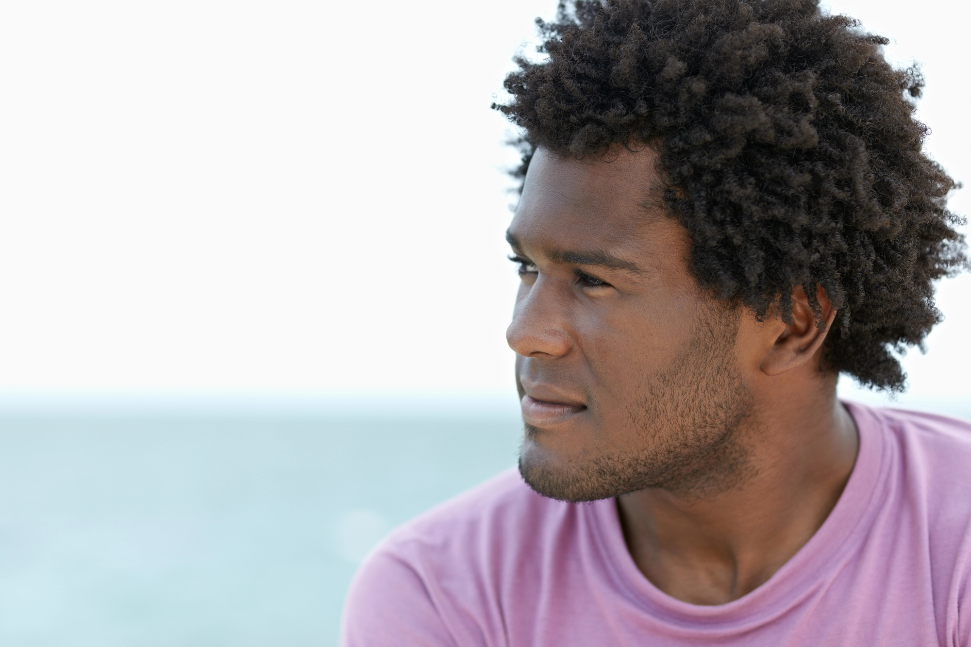 A person with short curly hair and a beard is gazing thoughtfully to the side. They are wearing a pink shirt and the background shows a blurred view of the ocean under a clear sky.