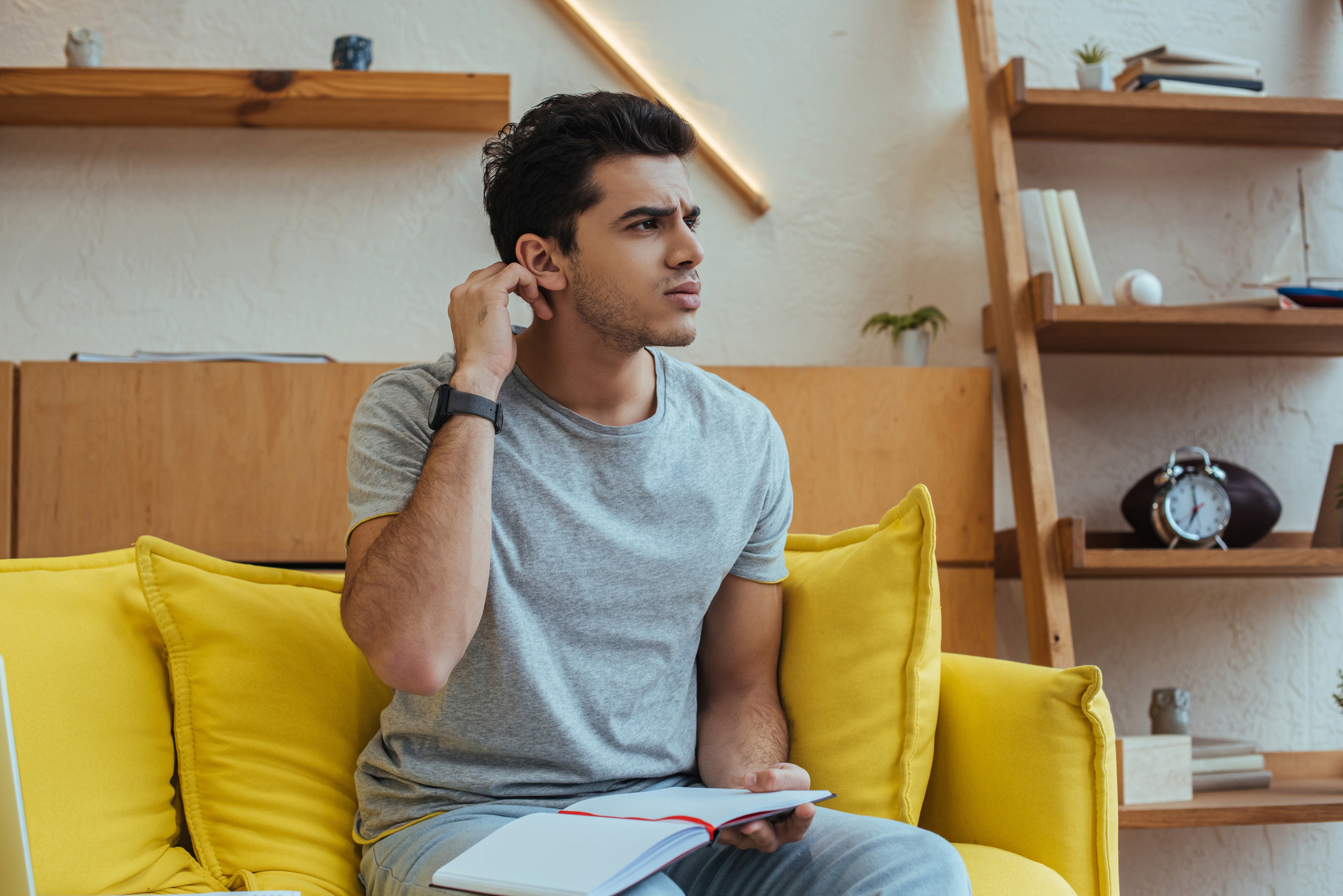 A young man sits on a yellow sofa in a living room, scratching his head and looking thoughtful. He holds an open notebook on his lap. The background includes shelves with books, a small plant, and a clock.