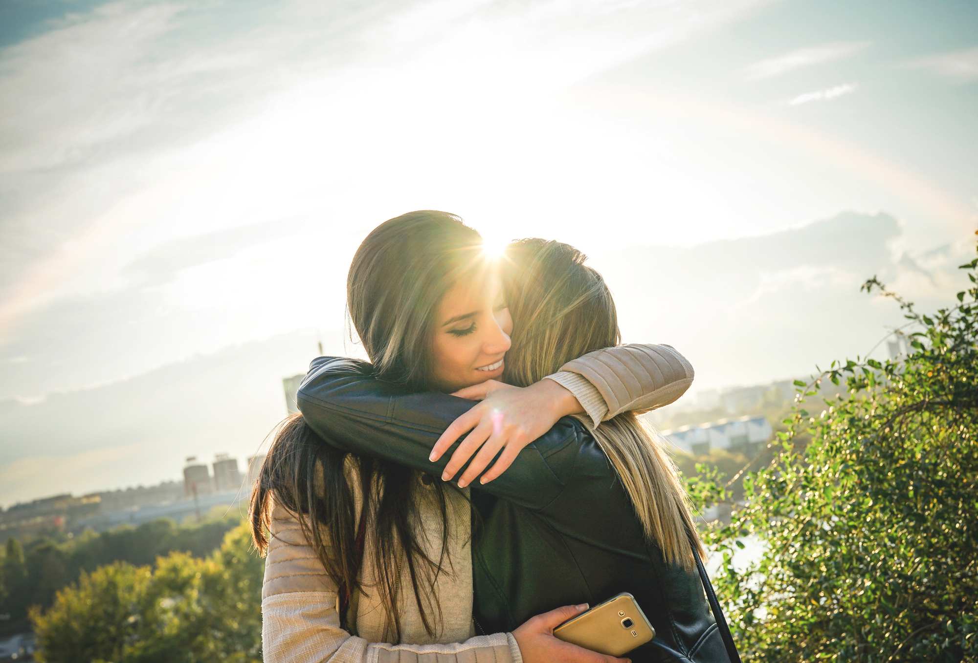Two women embrace on a sunny day, with a cityscape and greenery in the background. One woman, with long brown hair, is smiling. The other woman holds a smartphone in her hand. The sun is shining brightly, casting a warm glow and creating a partial rainbow in the sky.