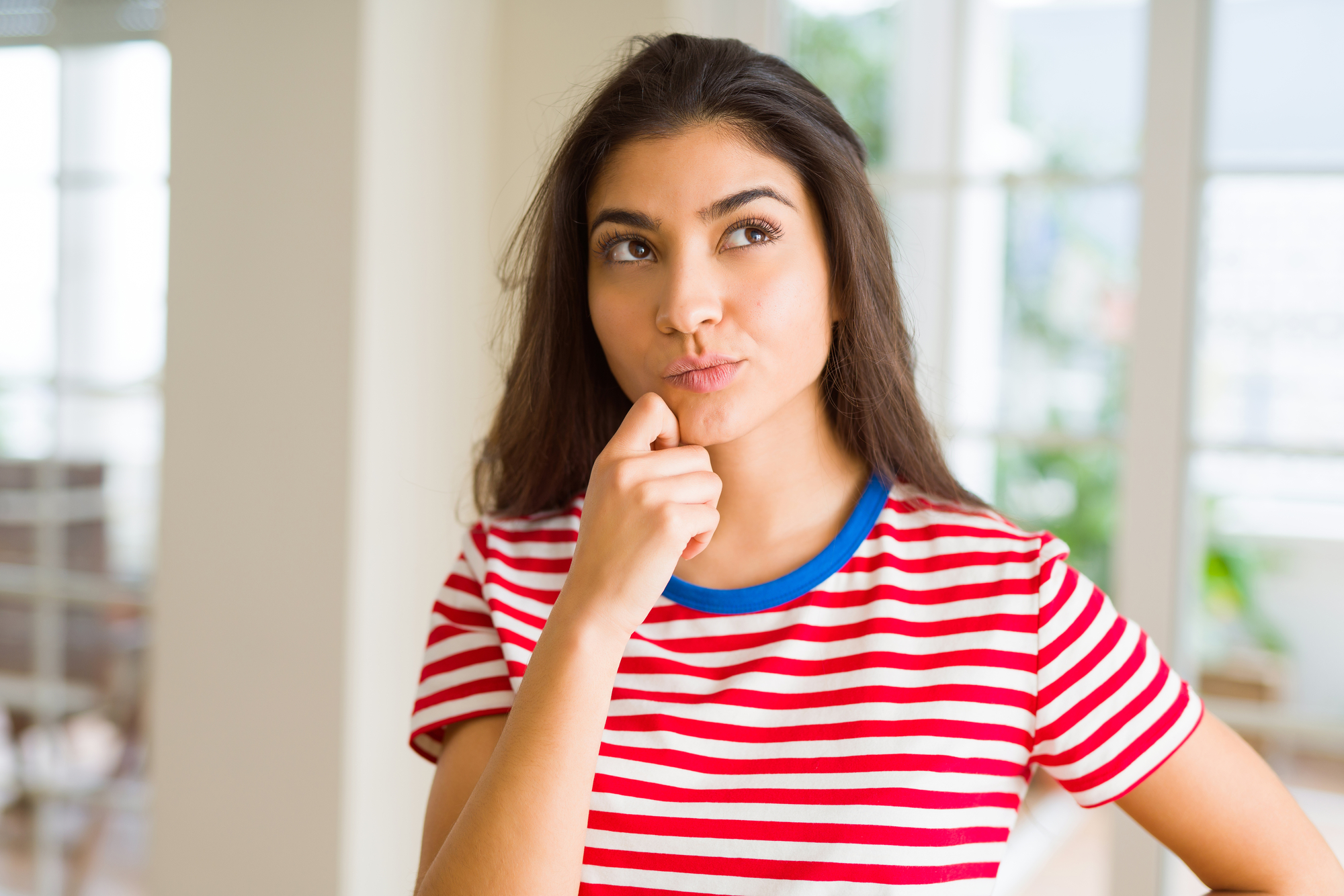 A woman with long dark hair wearing a red and white striped shirt is standing indoors, thoughtfully placing her hand on her chin. She looks upwards as if pondering something. The background is blurred, showing a well-lit room with windows.