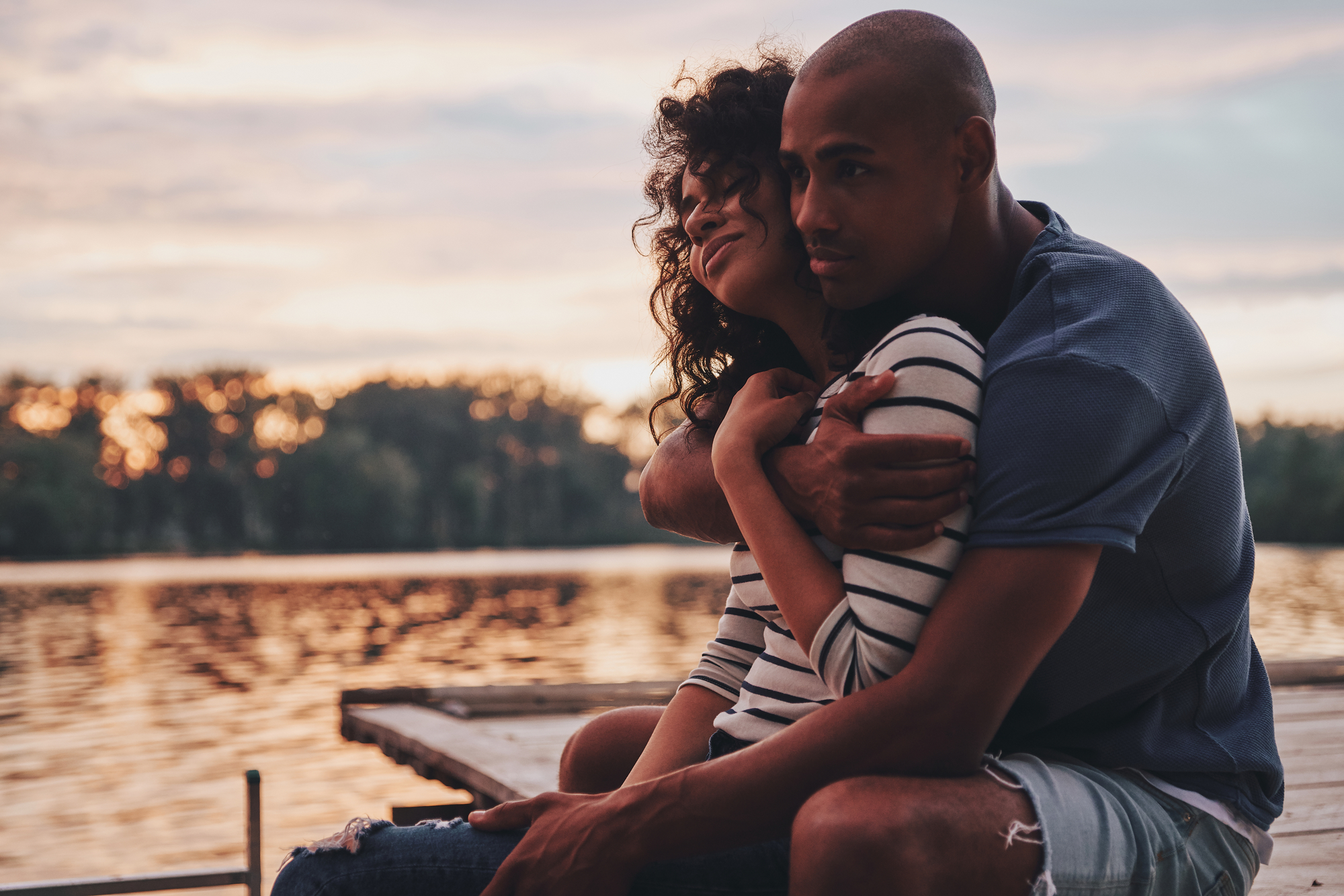 A couple sits on a dock overlooking a serene lake during sunset. The woman, wearing a striped shirt, is smiling while being embraced from behind by the man, who is wearing a blue shirt and shorts. The sky is painted with hues of orange and pink.