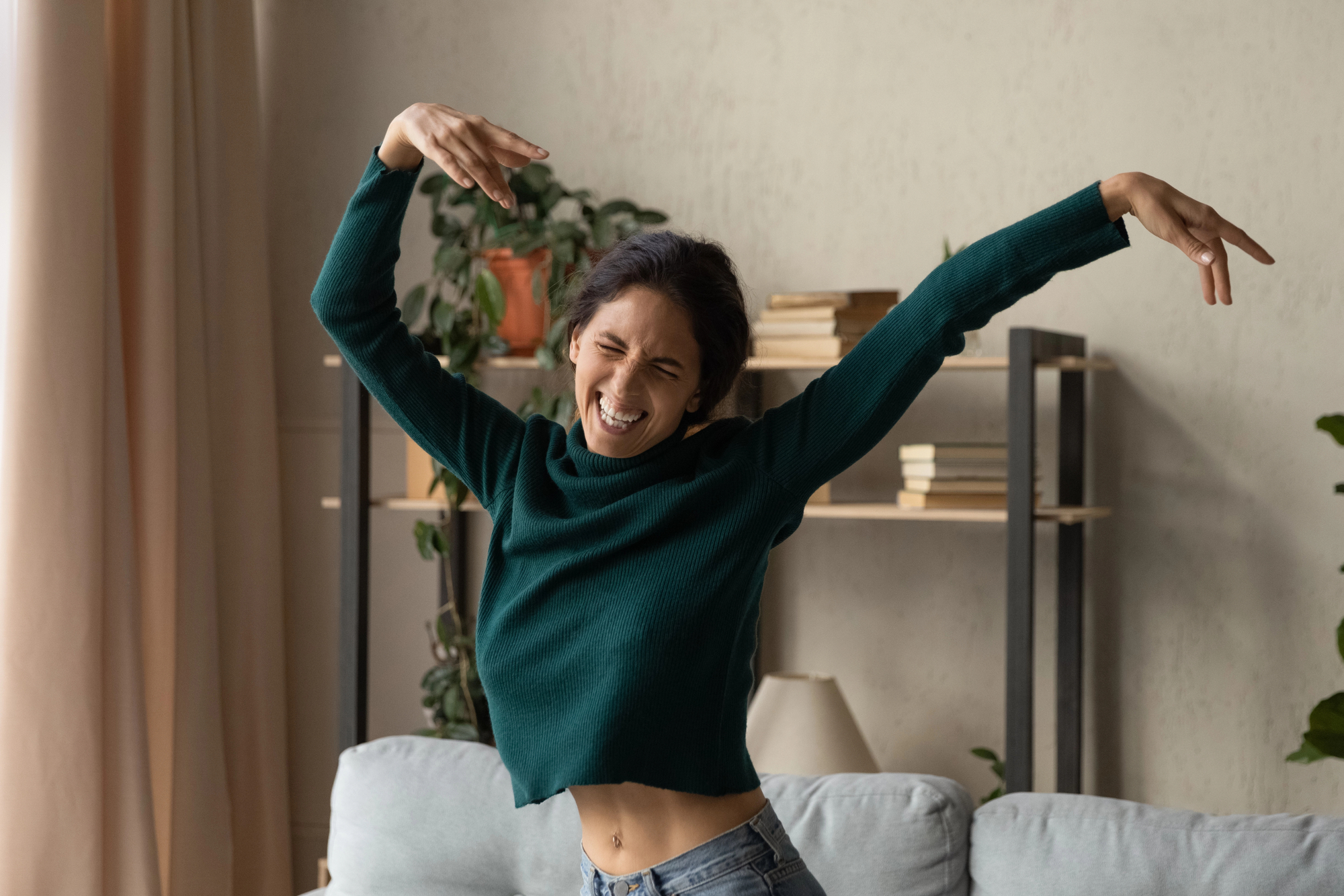 A woman wearing a green sweater is dancing with her eyes closed and smiling, her arms raised in a playful manner. She is standing in a living room with a soft couch and a bookshelf filled with books and plants in the background.